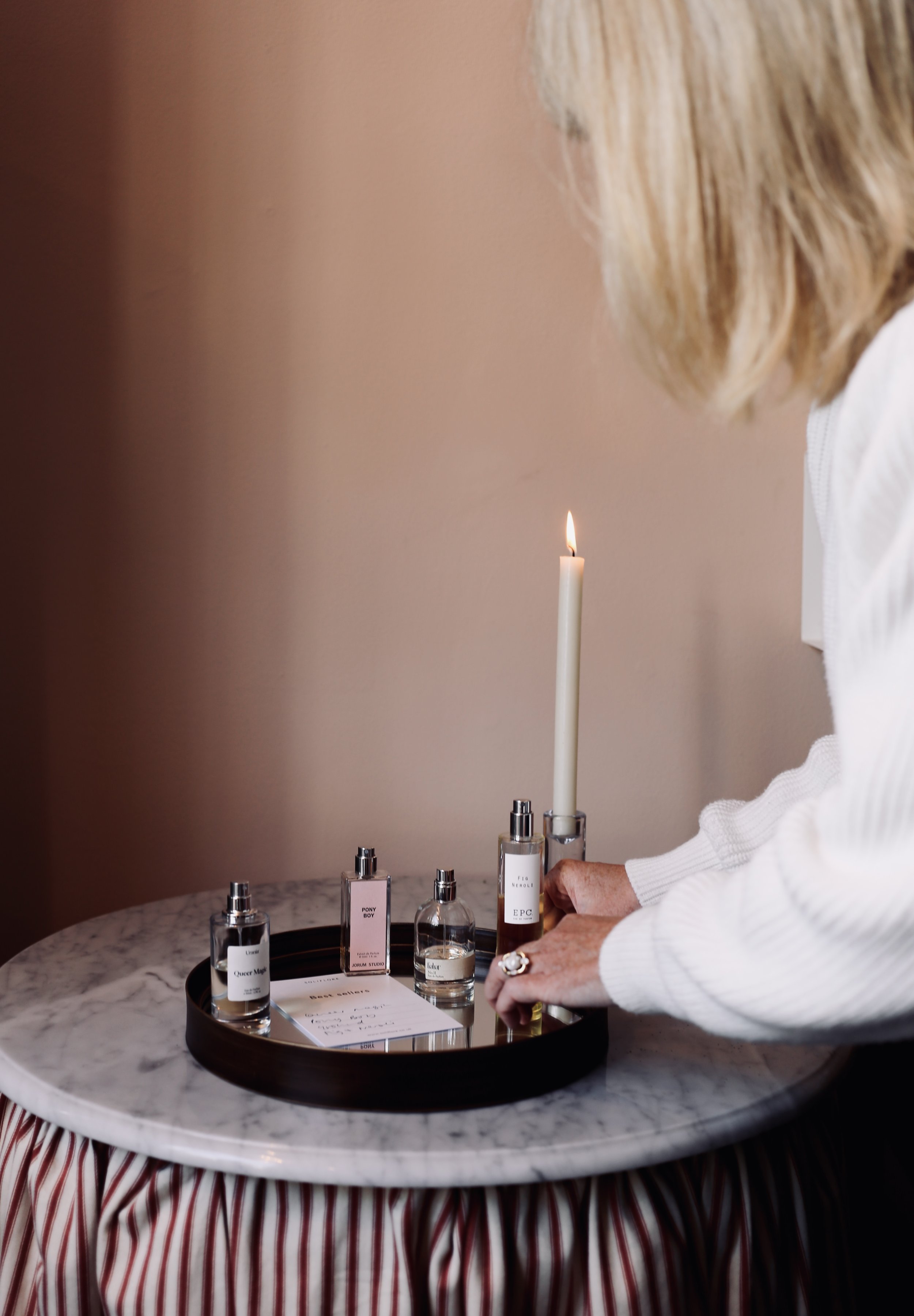 A woman with blonde hair picks up a small perfume bottle from a black tray on a marble table, with a lit white candle and several perfume bottles around it.