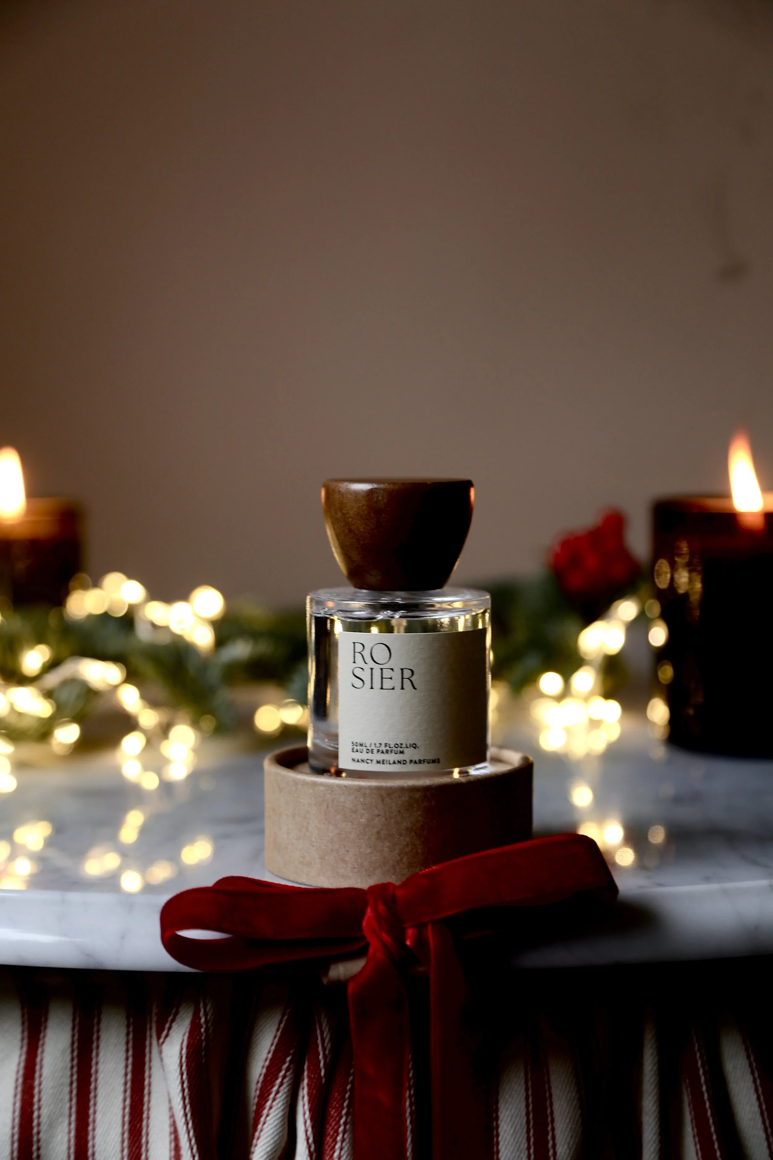 A perfume bottle labeled "Rosier" placed on a round cardboard box, decorated with a red ribbon, with holiday candles and lights in the background.