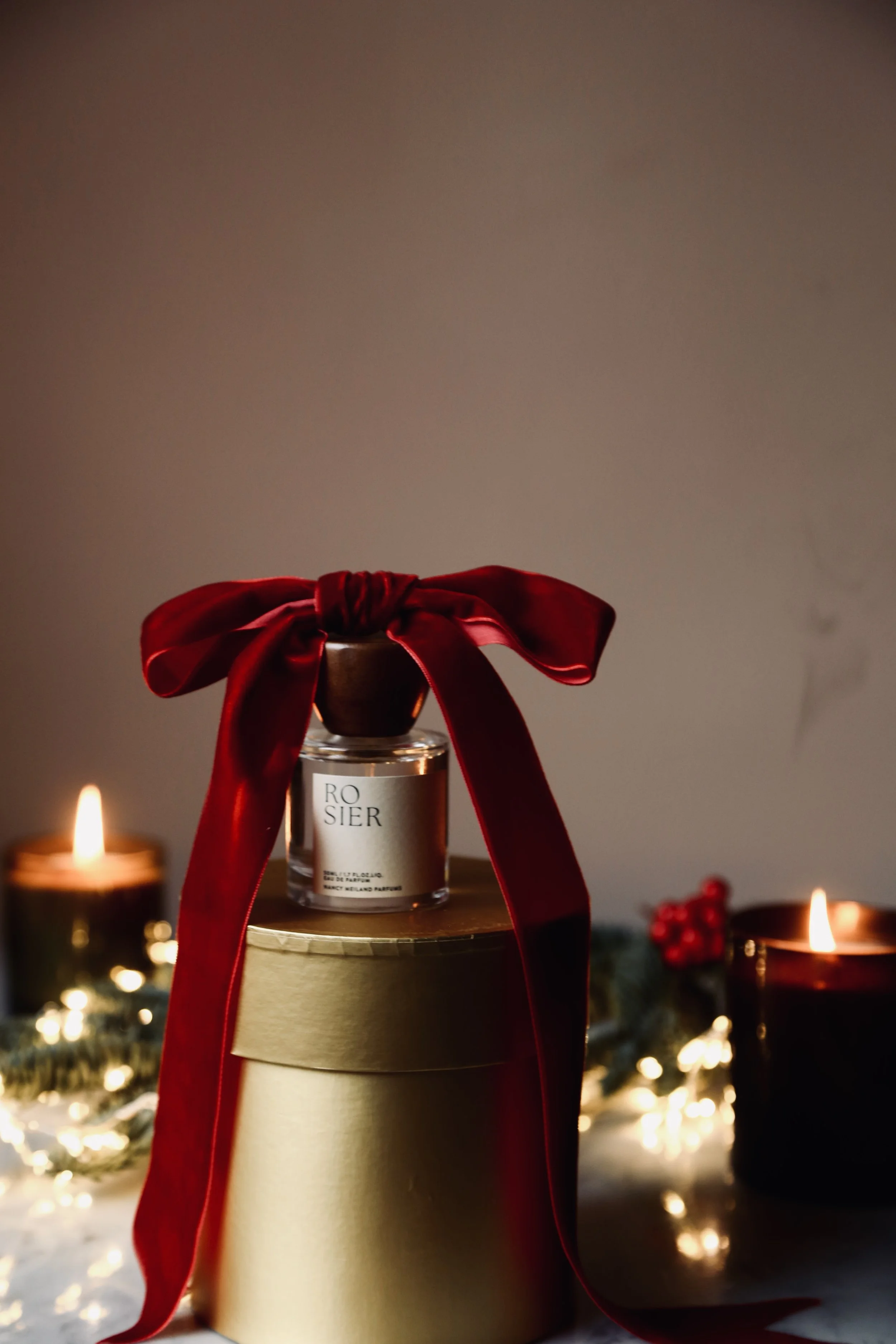 A perfume bottle with a red satin ribbon bow on top, placed on a cylindrical gold box, with Christmas candles and decorations in the background.