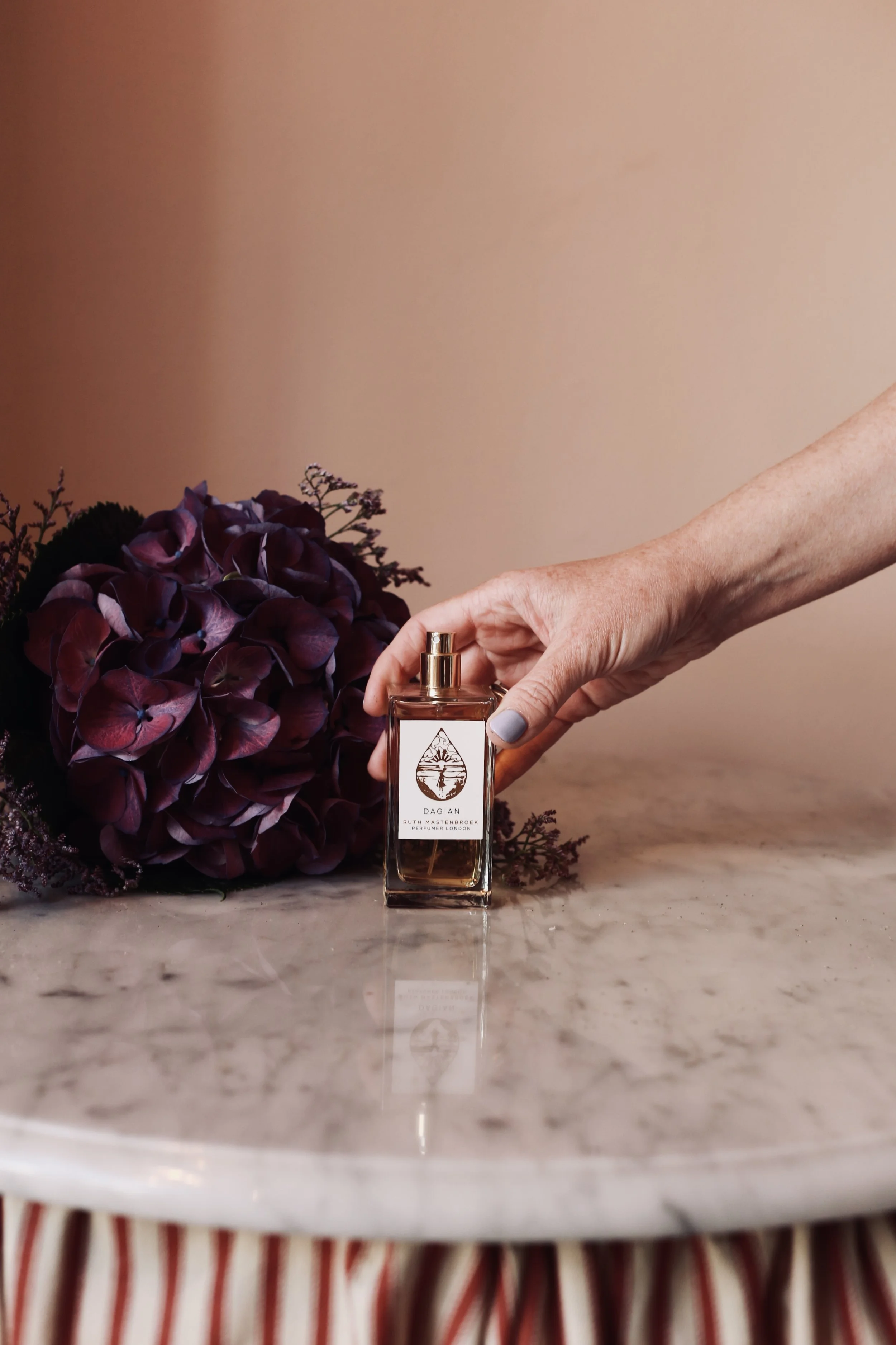 A hand with painted nails holding a perfume bottle labeled 'DAGIAN' and 'RUTH MASTENBROEK PERFUMER LONDON', placed on a marble surface with purple flowers nearby.