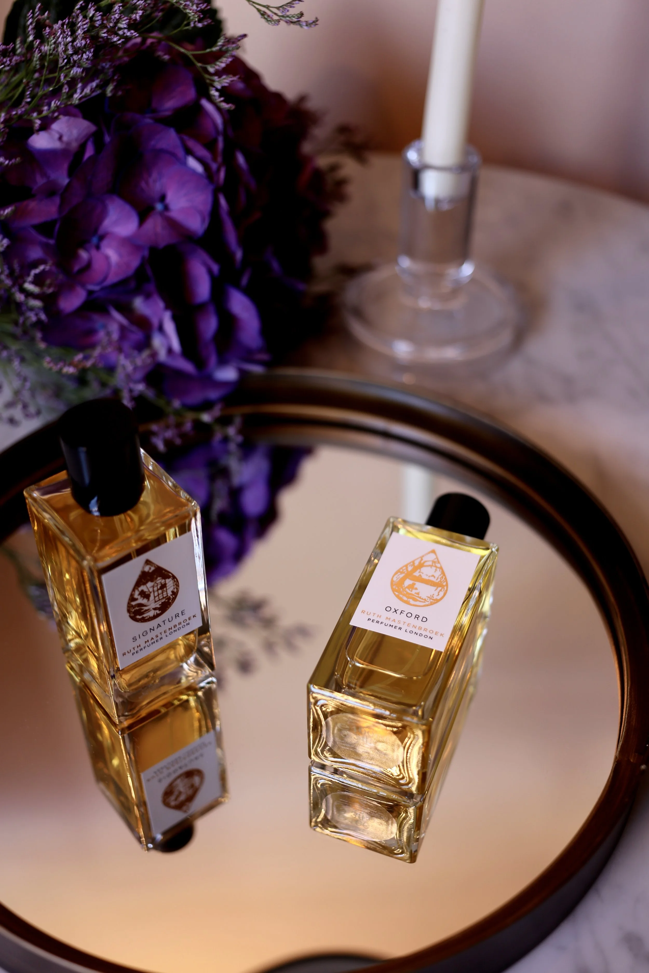 Two perfume bottles labeled 'Signature' and 'Oxford' placed on a reflective round tray, with a purple floral bouquet and a white candle in a glass holder in the background.