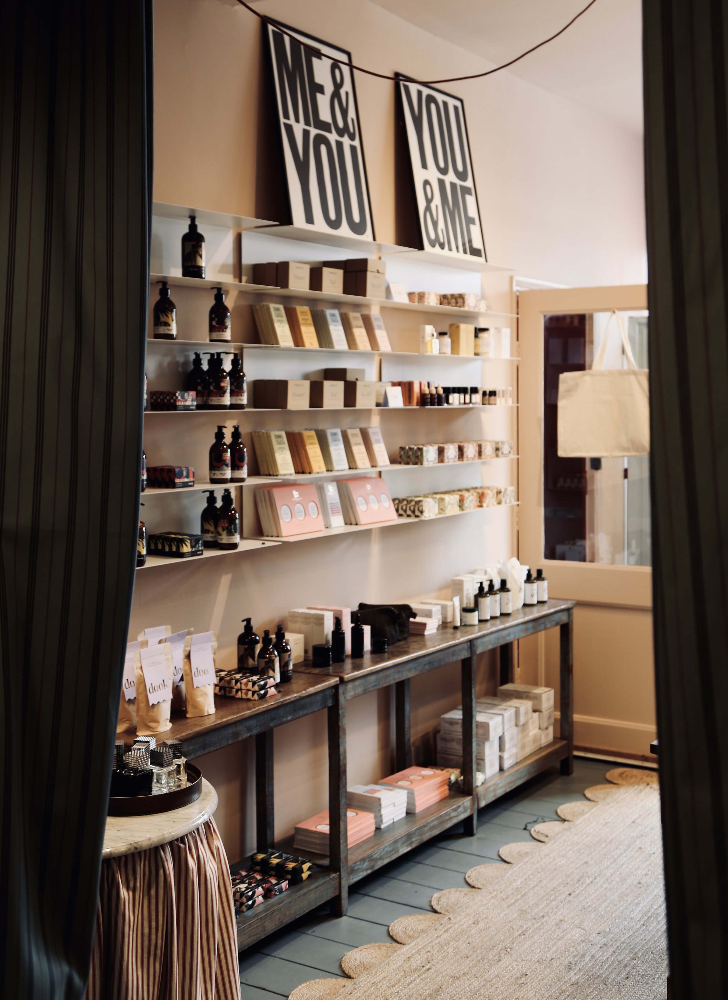 Interior of a boutique shop with shelves displaying various beauty or skincare products, including bottles, boxes, and packages. Signs hanging above read 'Me & You' and 'You & Me'.