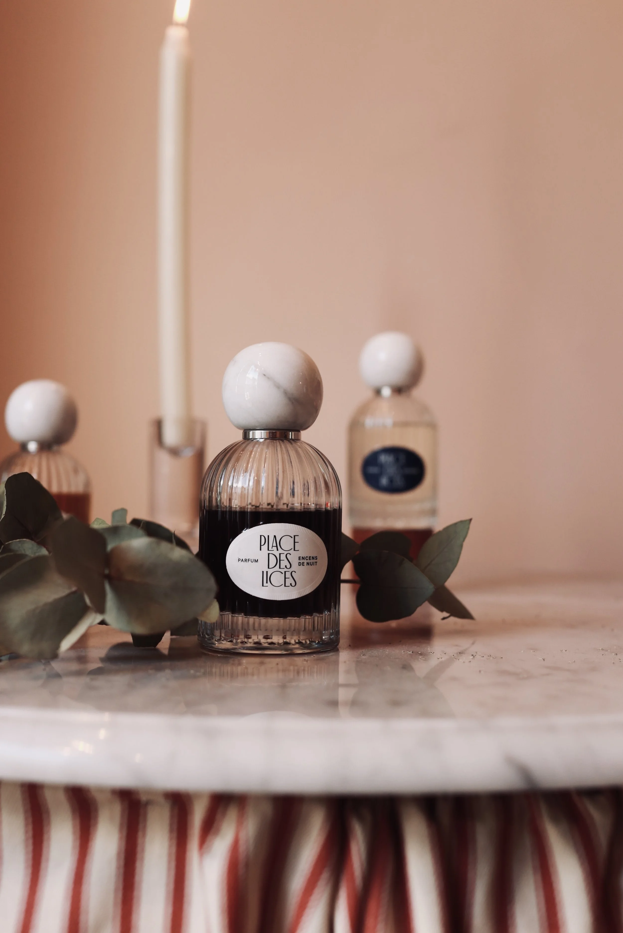 Perfume bottles with marble caps and a lit candle on a marble surface, decorated with green leaves, against a pink background.
