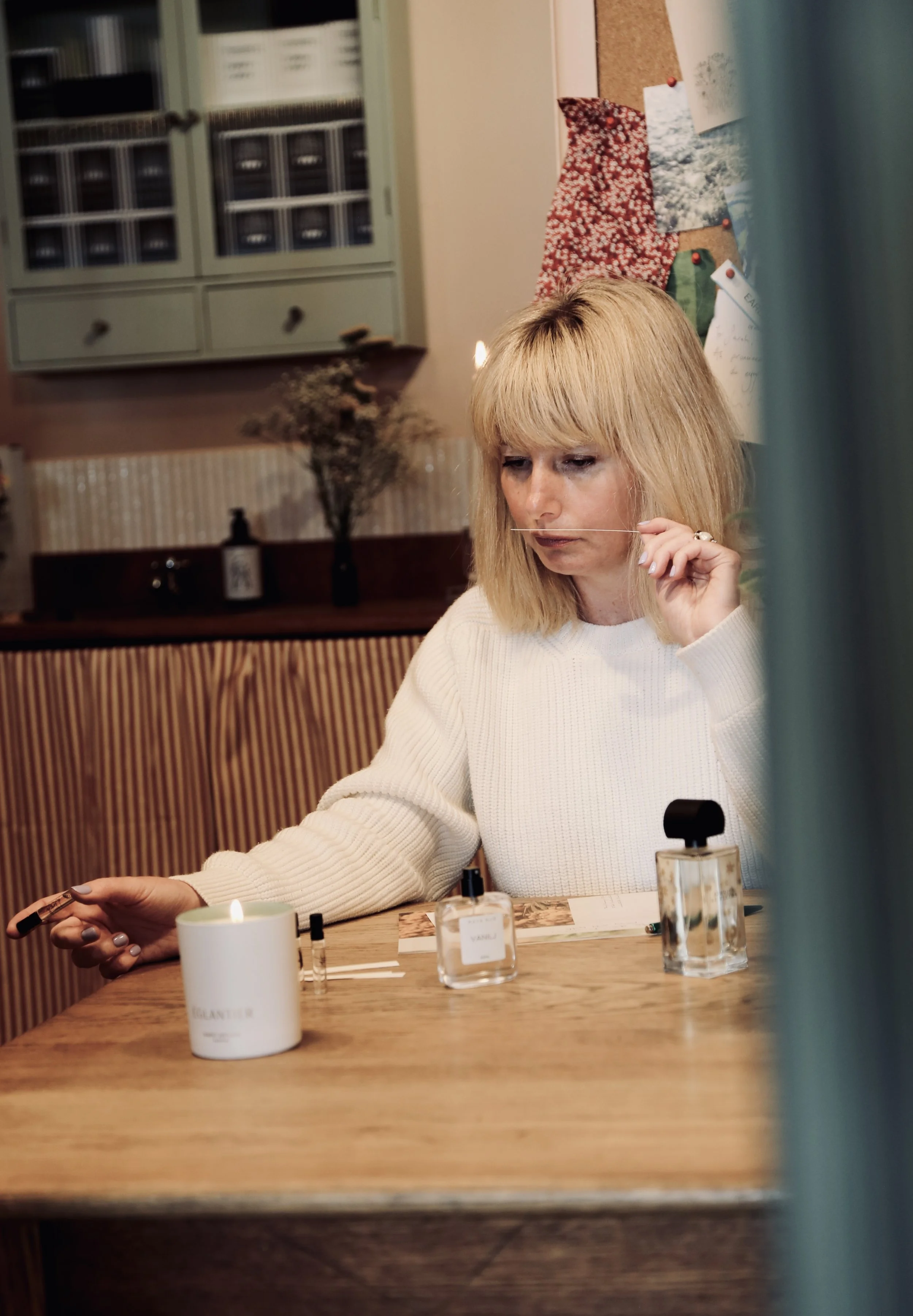 A woman with blonde hair is at a table, testing perfume scent strips, with perfume bottles and a candle on the table. She is wearing a white sweater and is in a cozy, decorated room.