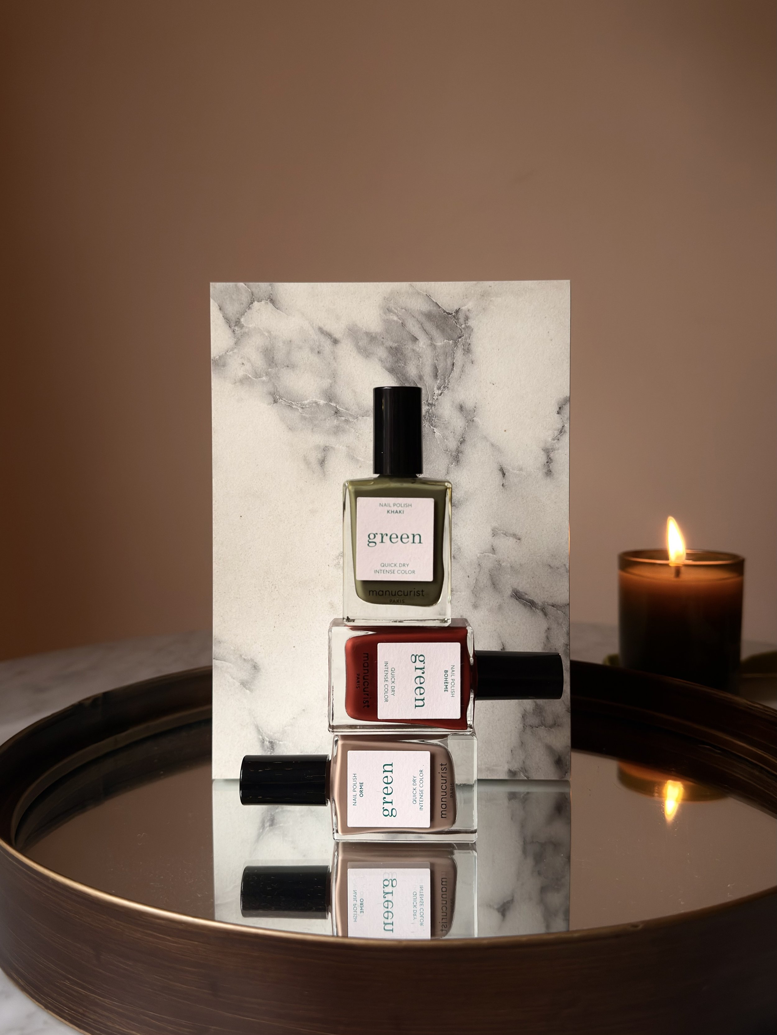 Three bottles of nail polish labeled "green," stacked vertically, with reflection on a mirror, set against a marble background, with a lit candle on a holder in the background.
