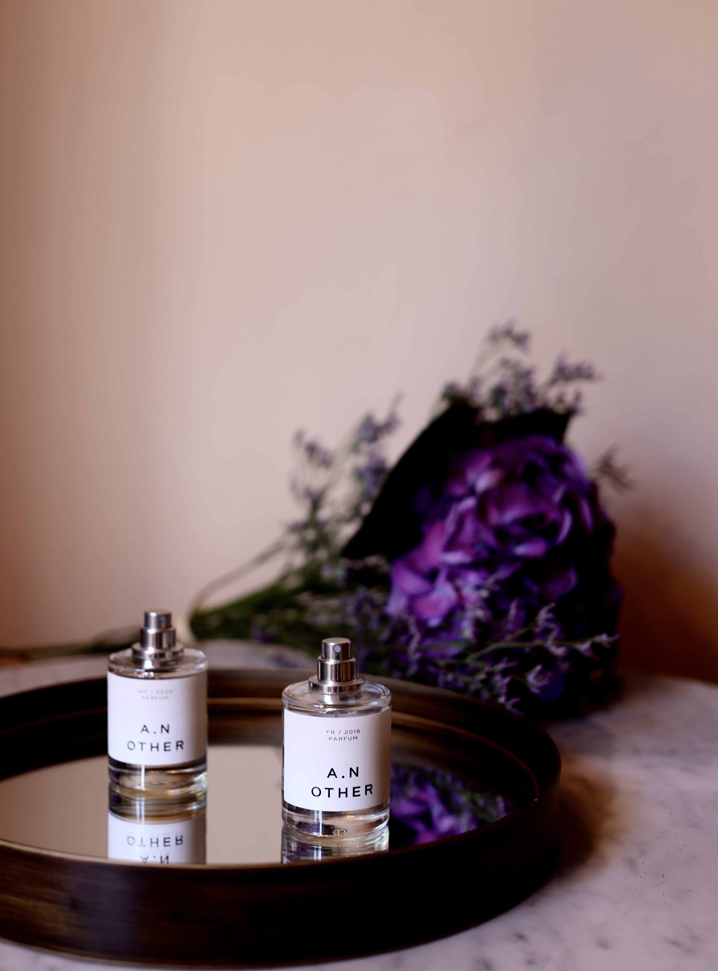 Two perfume bottles labeled "A.N OTHER" are reflected on a small round mirror, with a bouquet of purple flowers in the blurred background.