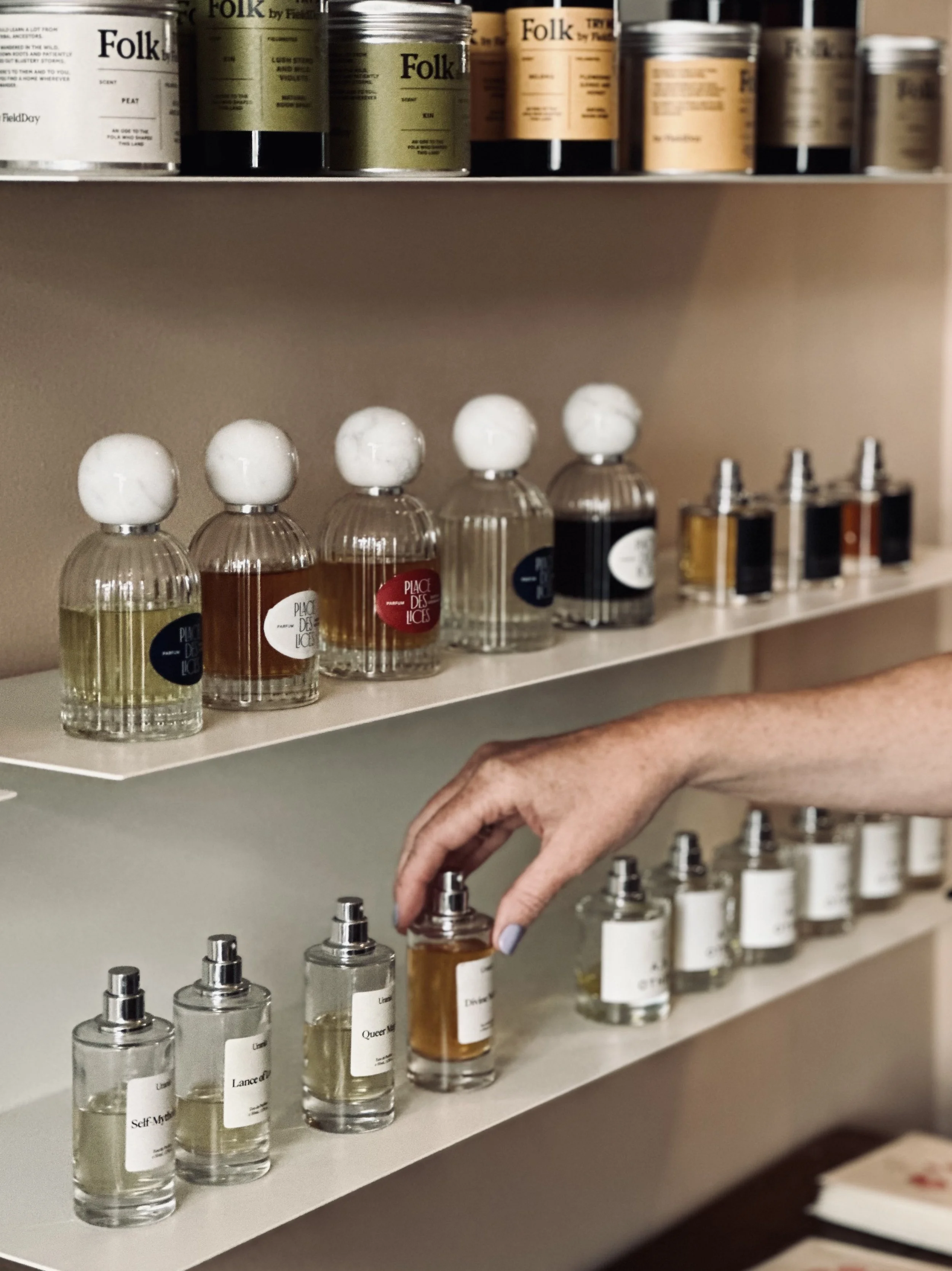 A hand reaching for a perfume bottle on a shelf filled with various perfume bottles and fragrance oils.