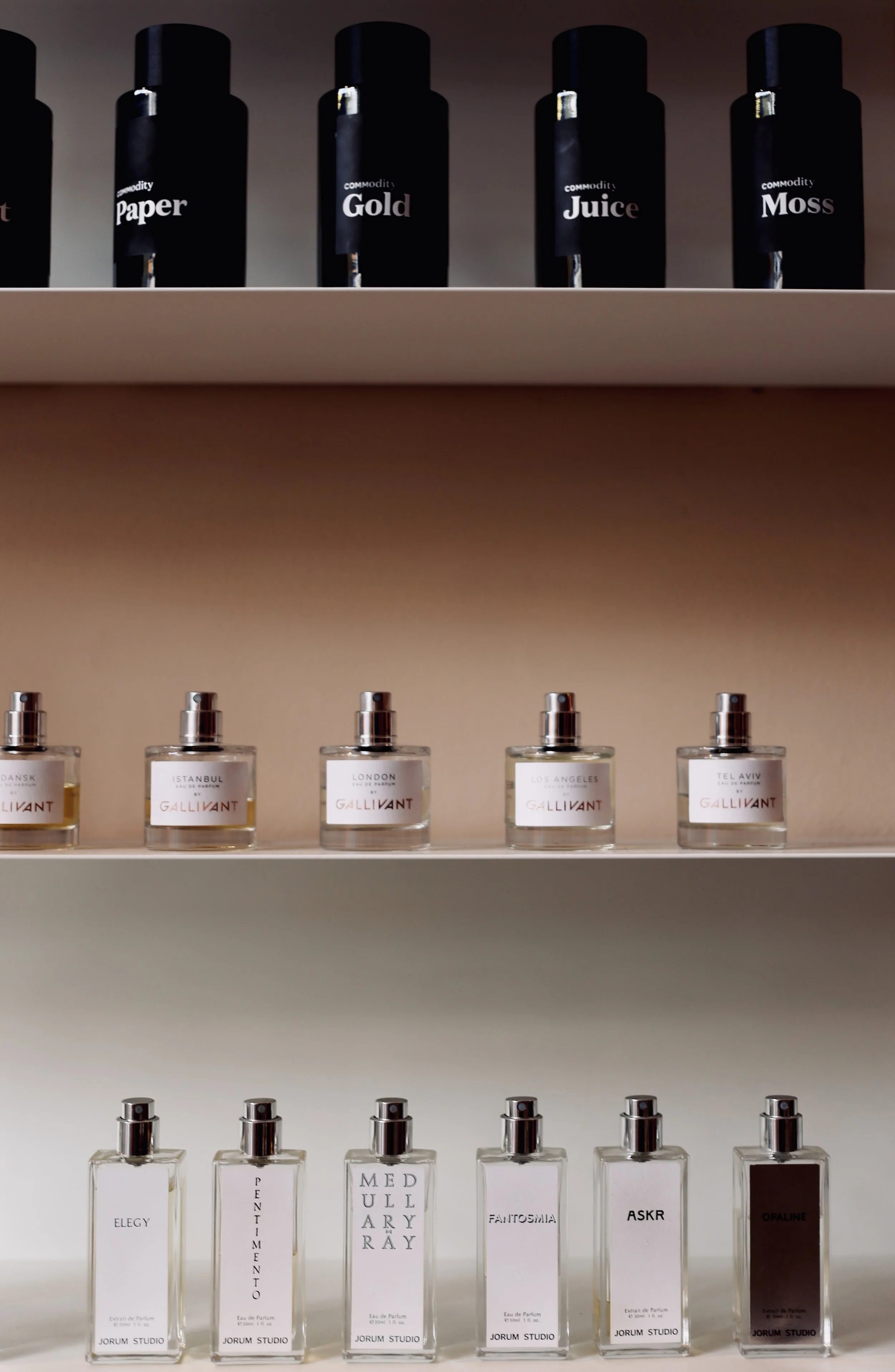  shelves with black bottles labeled 'Commodity Paper,' 'Commodity Gold,' 'Commodity Juice,' 'Commodity Moss' on the top shelf; smaller perfume bottles labeled with various names on the middle and bottom shelves.