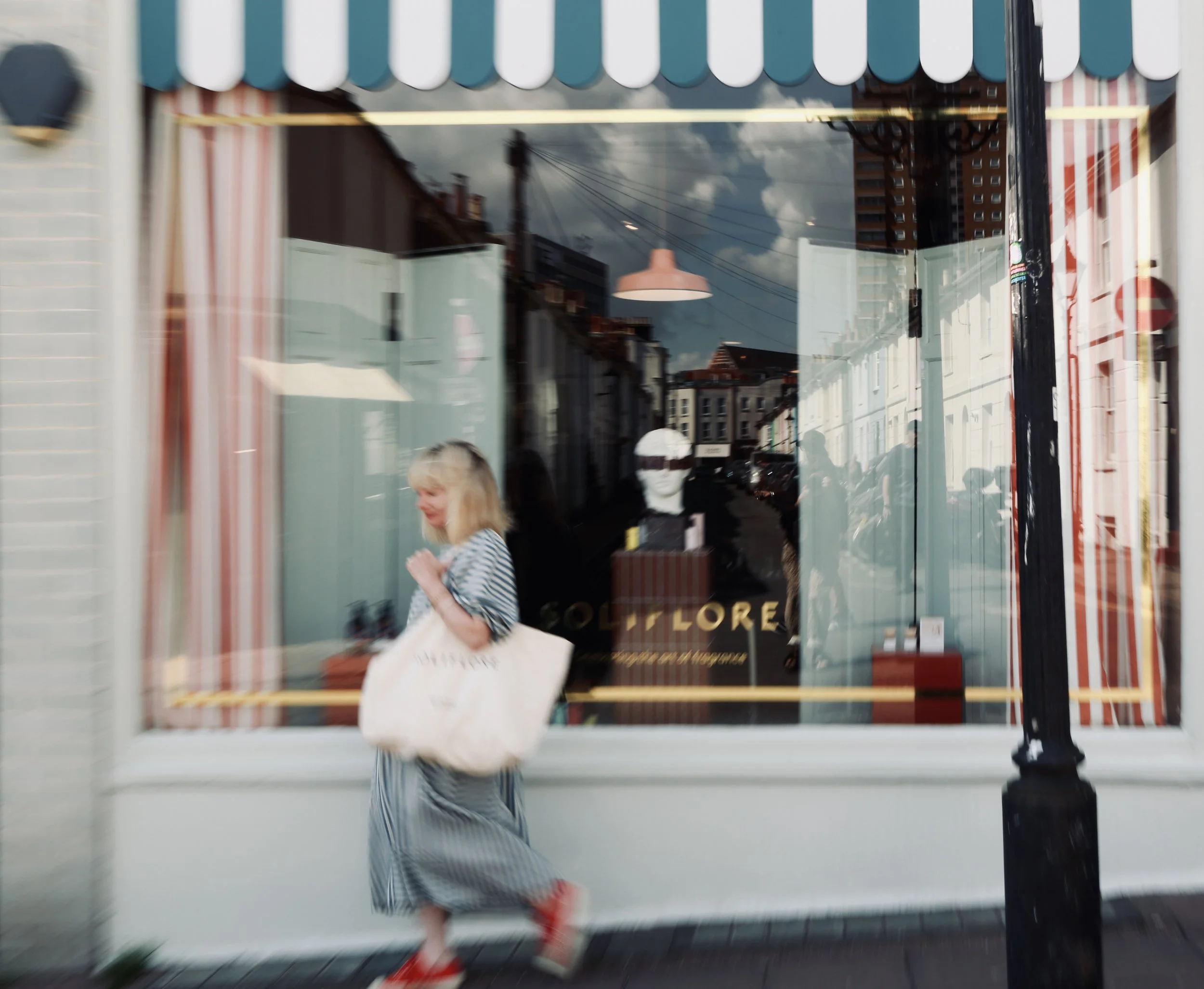 Blurred photo of a woman walking past a storefront window with striped curtains, a mannequin head with sunglasses inside the shop, and a reflection of buildings and power lines outside.