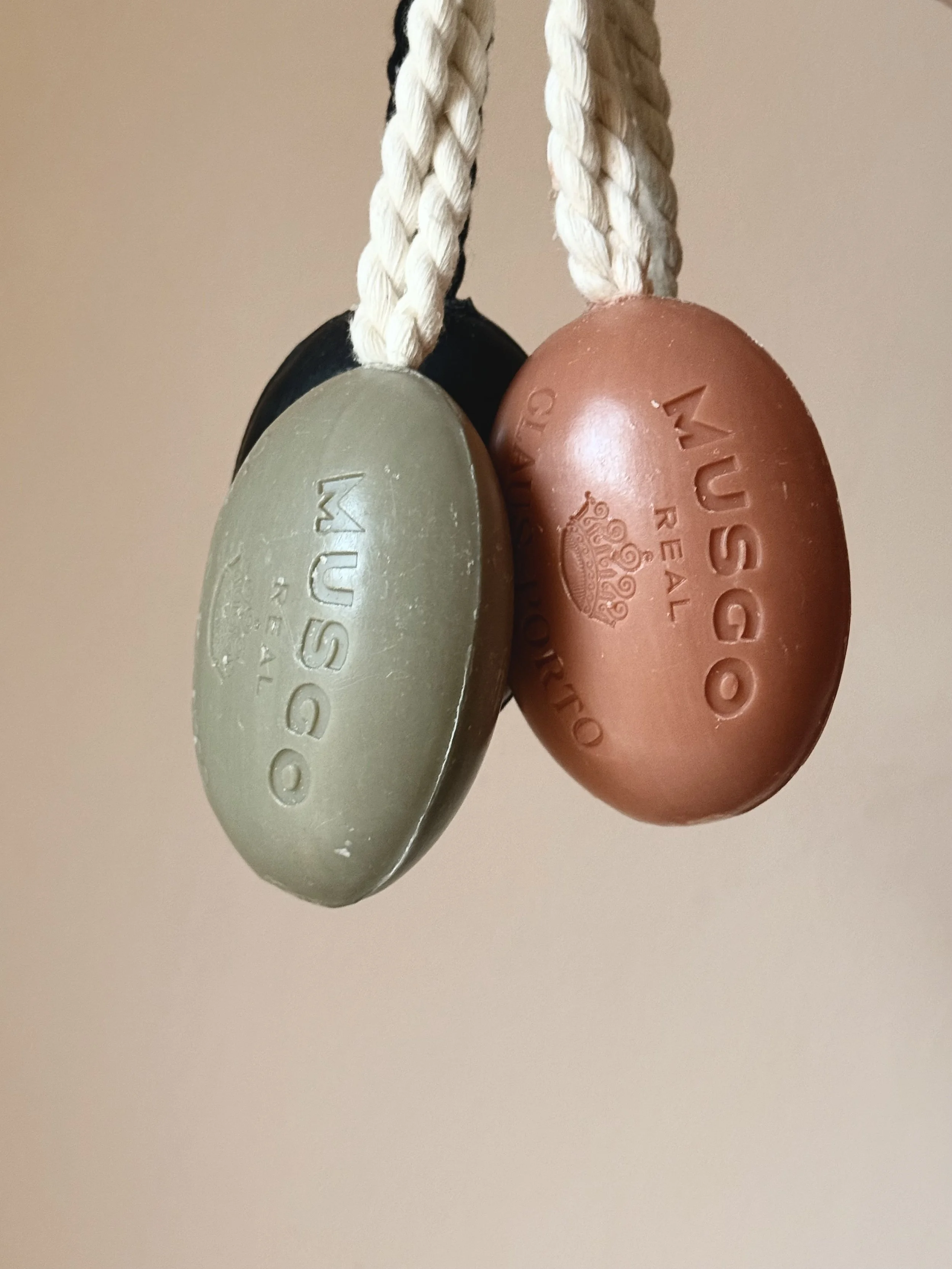 Three oval-shaped MUGO soap bars hanging on braided cords; one beige, one black, one brown, with the MUGO brand and crown logo embossed on each.