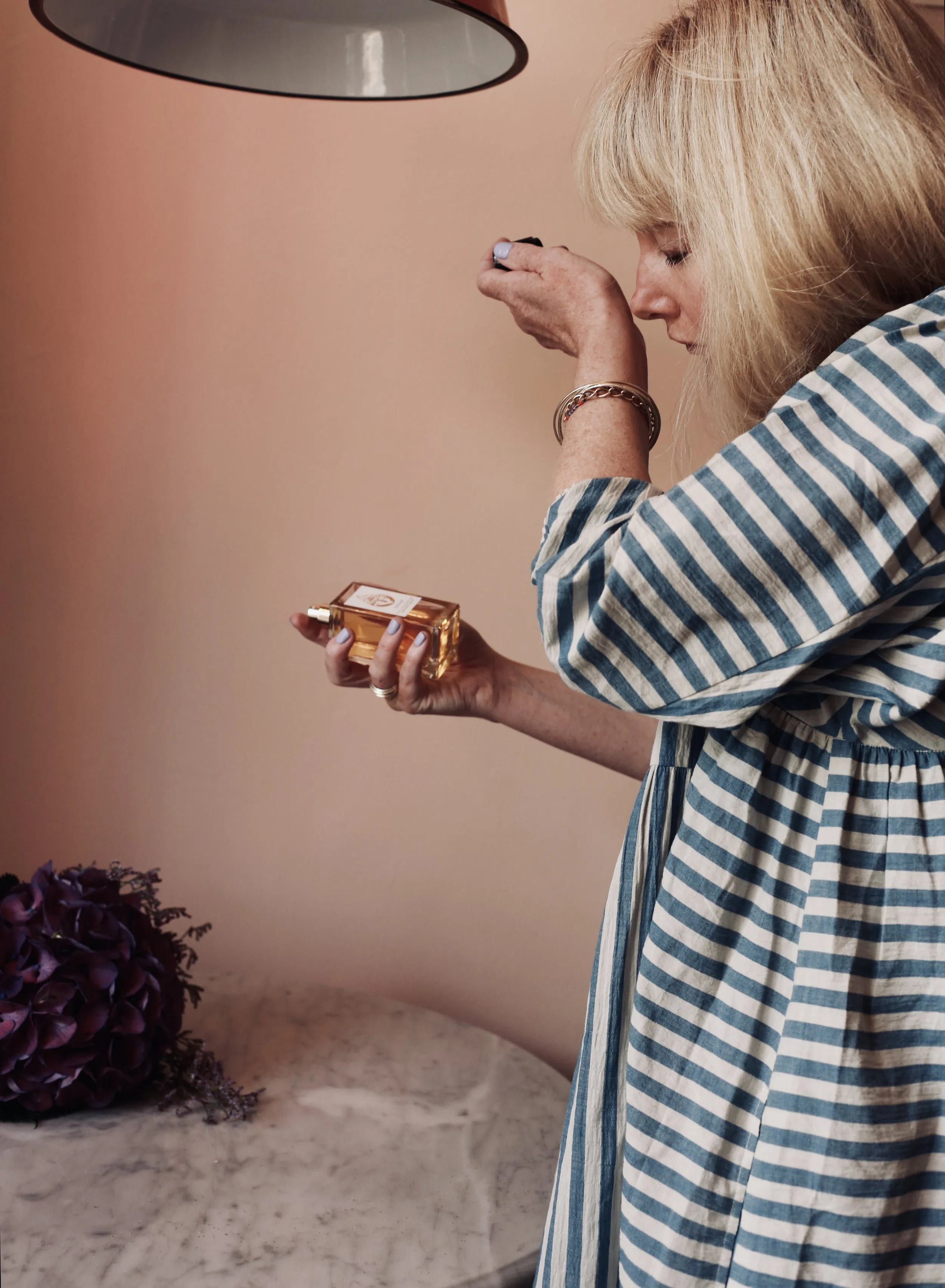 A woman with blonde hair wearing a blue and white striped dress is holding a perfume bottle and spraying perfume on herself in a room with pink walls, a hanging lamp, and purple flowers on a marble surface.