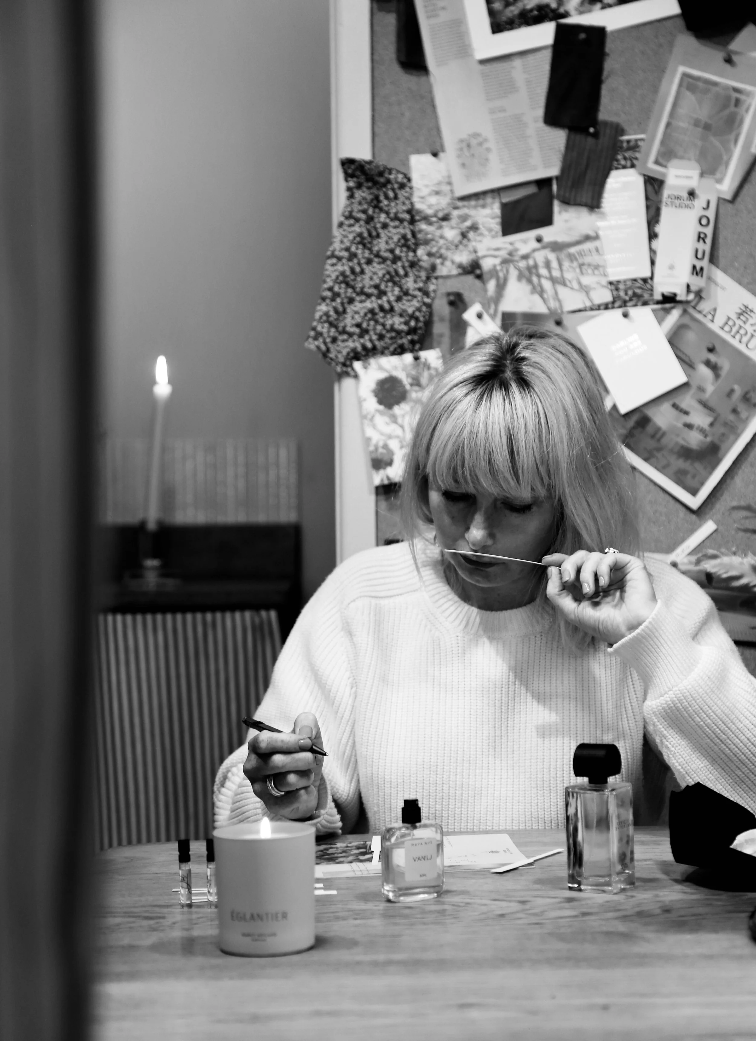 A woman with blonde hair seated at a desk, smelling a bottle of perfume with assorted perfume bottles and a lit candle in front of her, in a room with a bulletin board covered with various photos, papers, and fabric samples.