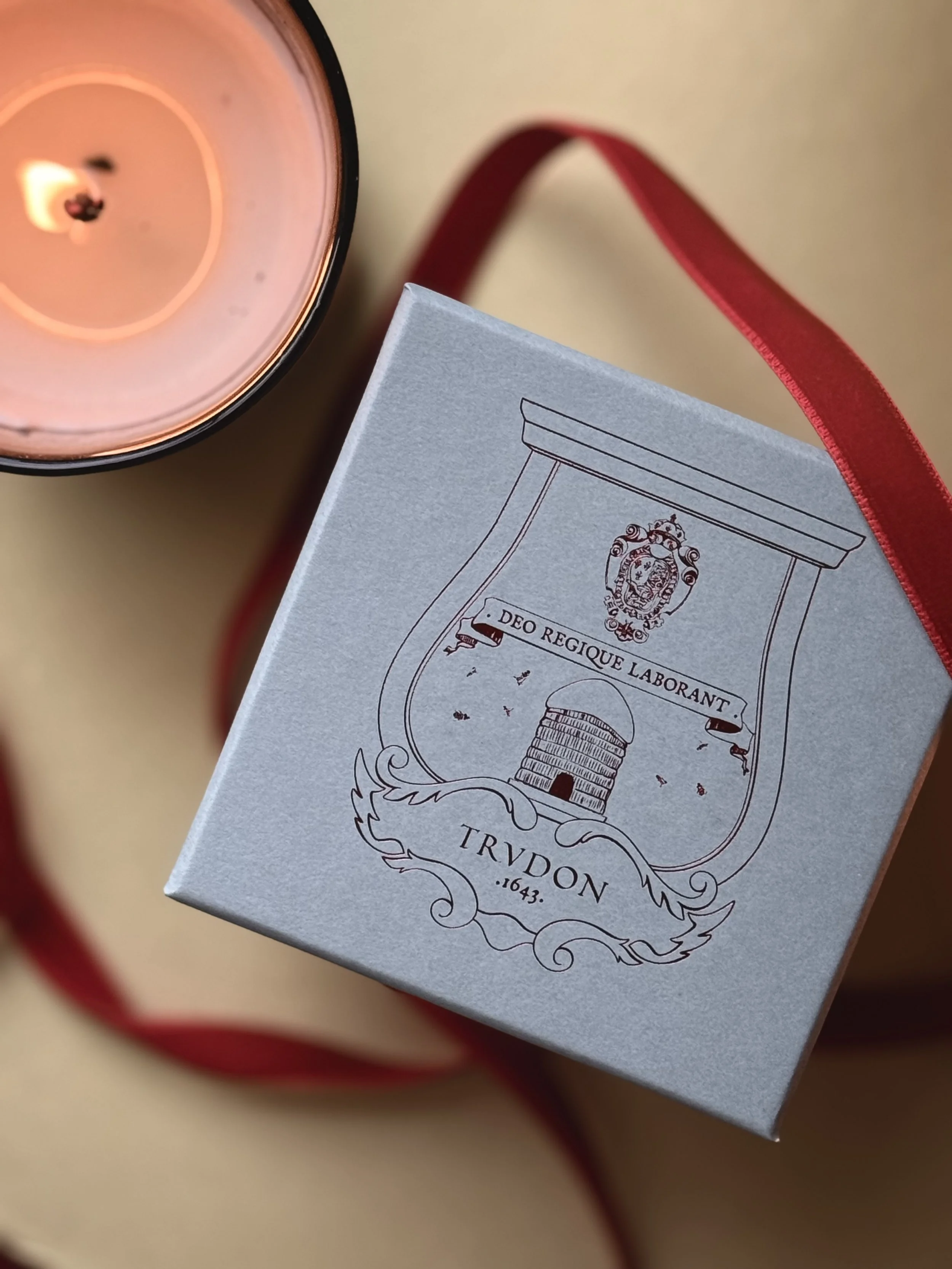 A close-up of a blue box with intricate line art and Latin text, alongside a lit candle in a clear container with a red ribbon around it.