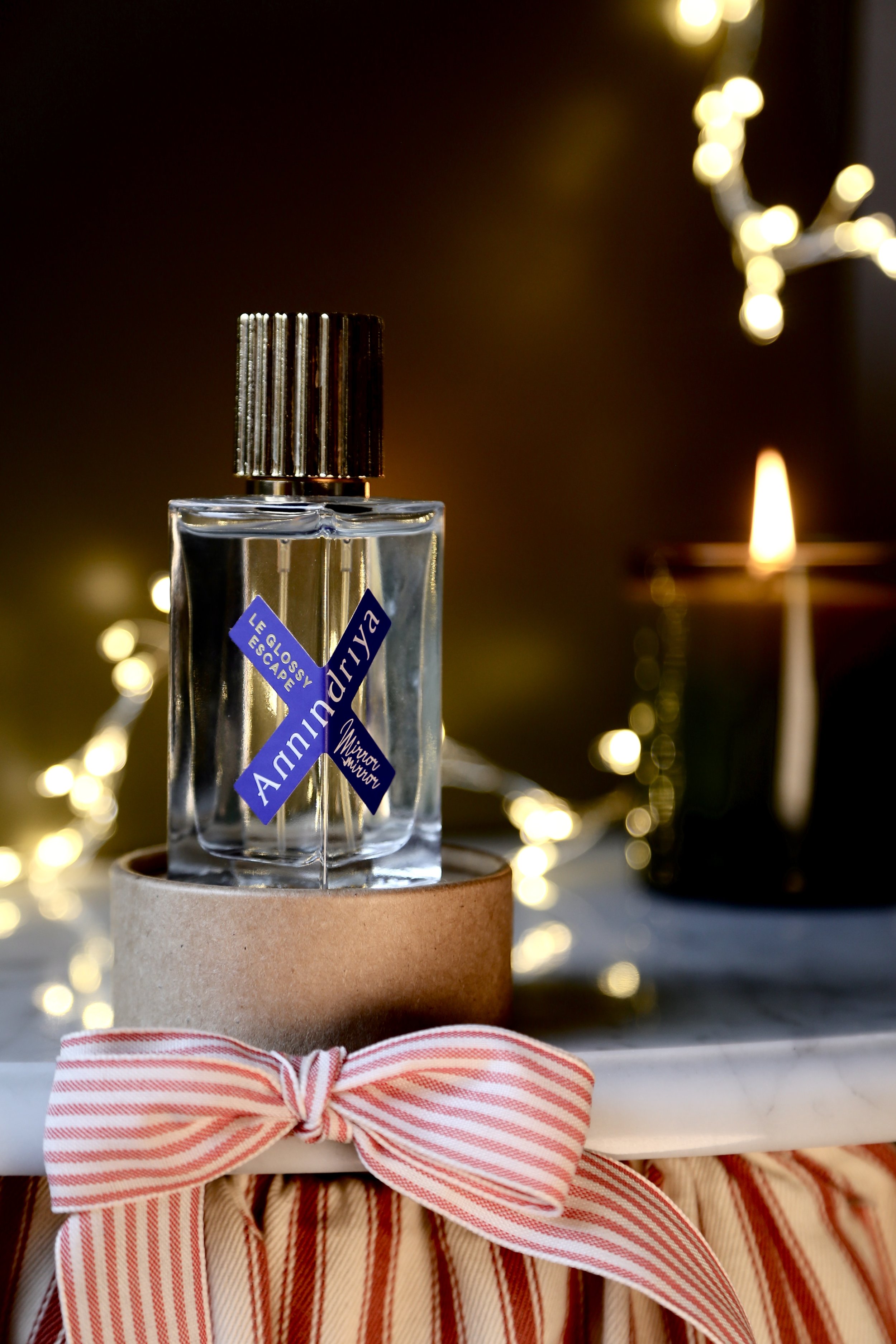 A perfume bottle labeled 'Amnudy,' placed on a round cardboard stand with a red and white striped ribbon tied around it. In the background, warm bokeh lights and a candle are visible.