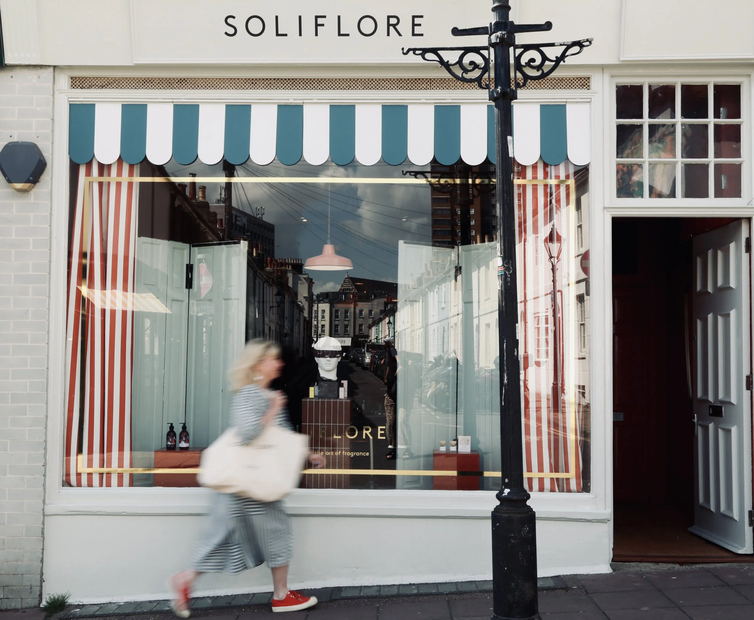 A storefront with a glass window displaying striped curtains, a sign that reads 'SOLIFLOR,' and a reflection of urban buildings and power lines. A woman with blonde hair, wearing a striped dress and red shoes, walks past, holding a large beige bag.