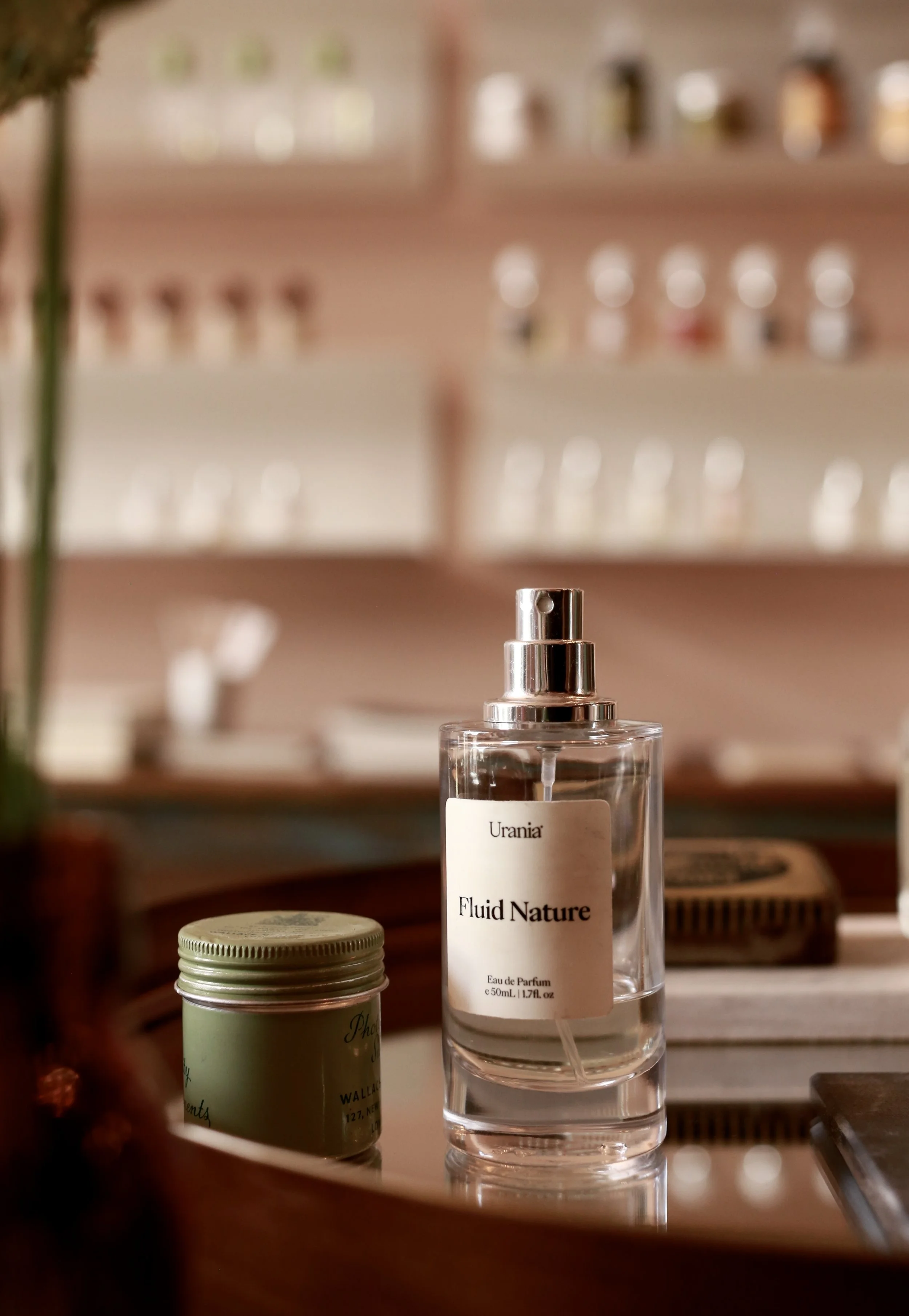 A clear perfume bottle labeled "Urania Fluid Nature" on a reflective surface, with a small green jar to its side, and a blurred background of shelves with bottles.