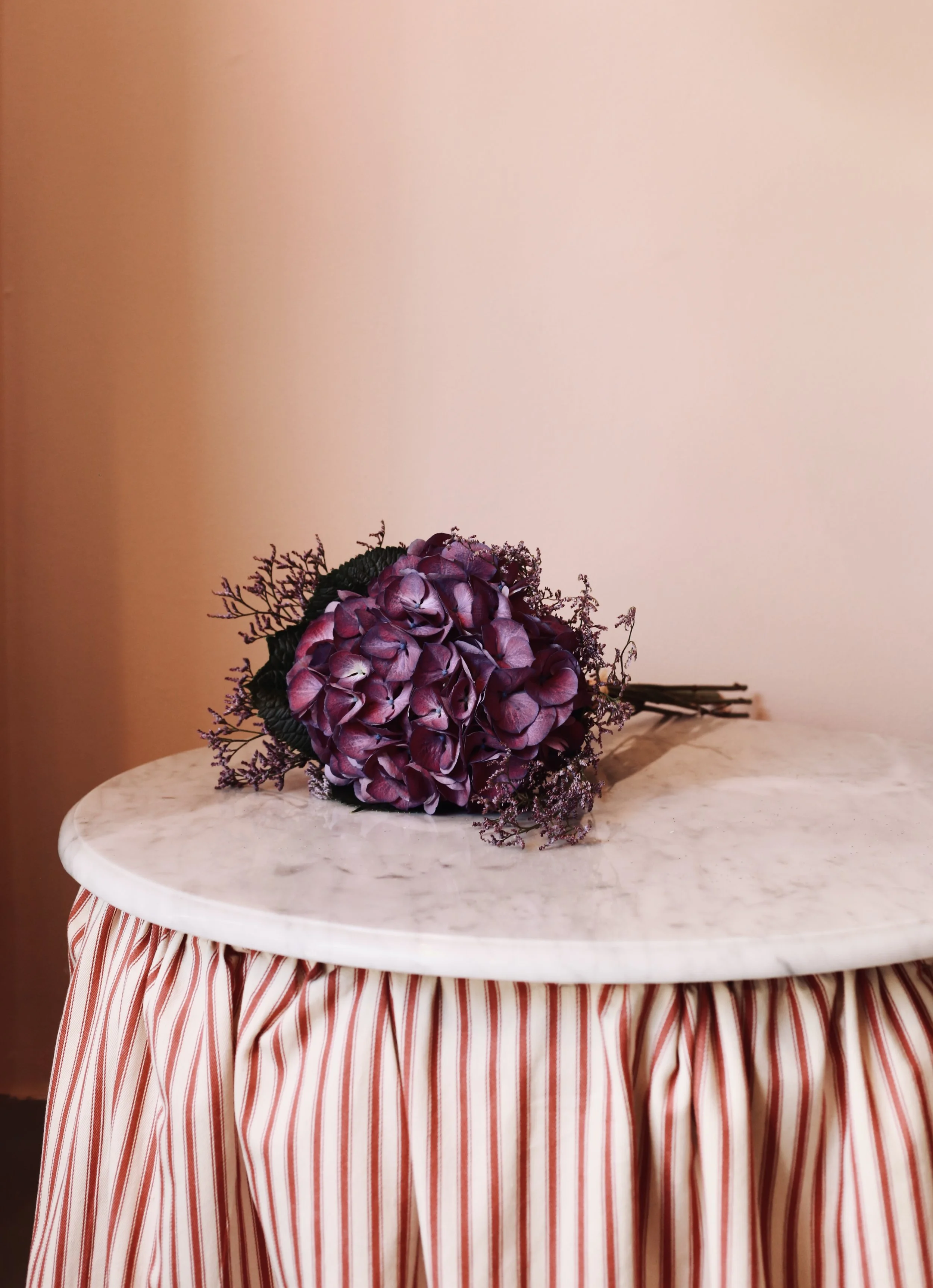 A bouquet of purple and pink hydrangeas with greenery, resting on a small round table with a marble top and striped fabric skirt, against a plain beige wall.