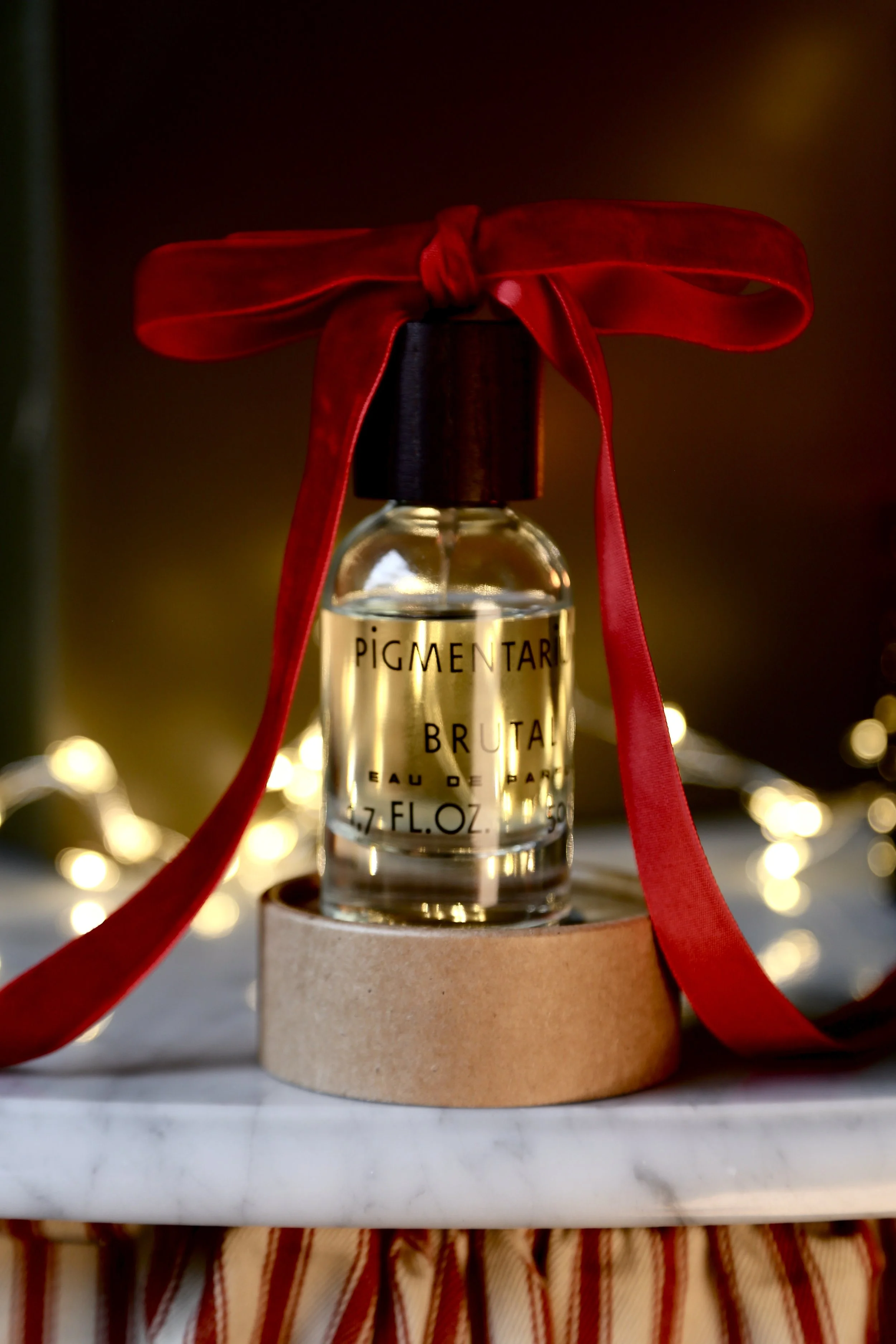 Clear glass bottle with perfume labeled 'Pigmentari Bruta Eau de Parfum' topped with a black cap and tied with a red ribbon, placed on a circular cardboard holder with blurred string lights in the background.