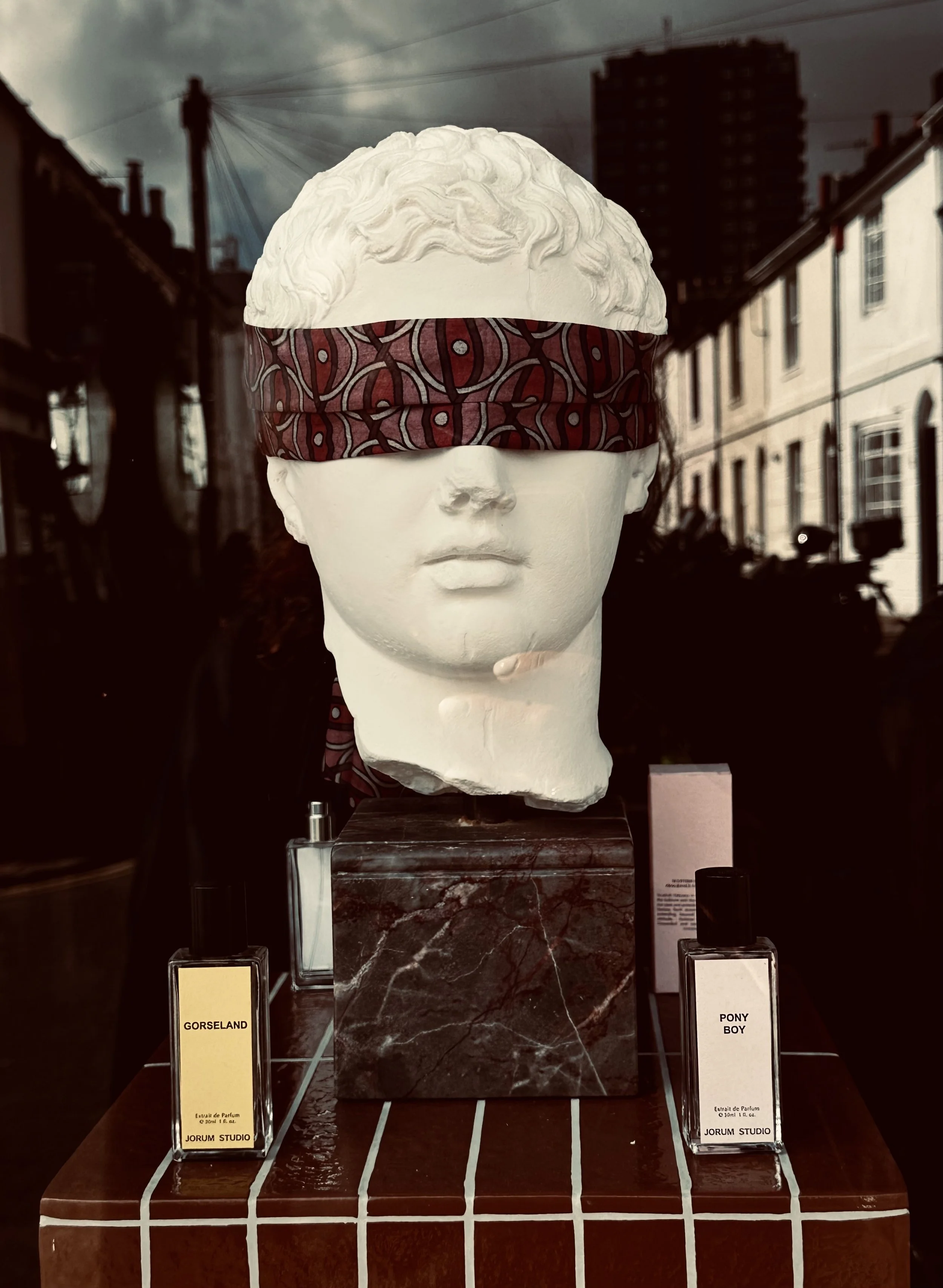 A mannequin head with curly hair, blindfolded with a patterned cloth, displayed in a shop window. The display includes perfume bottles labeled "Gorseland" and "Pony Boy."