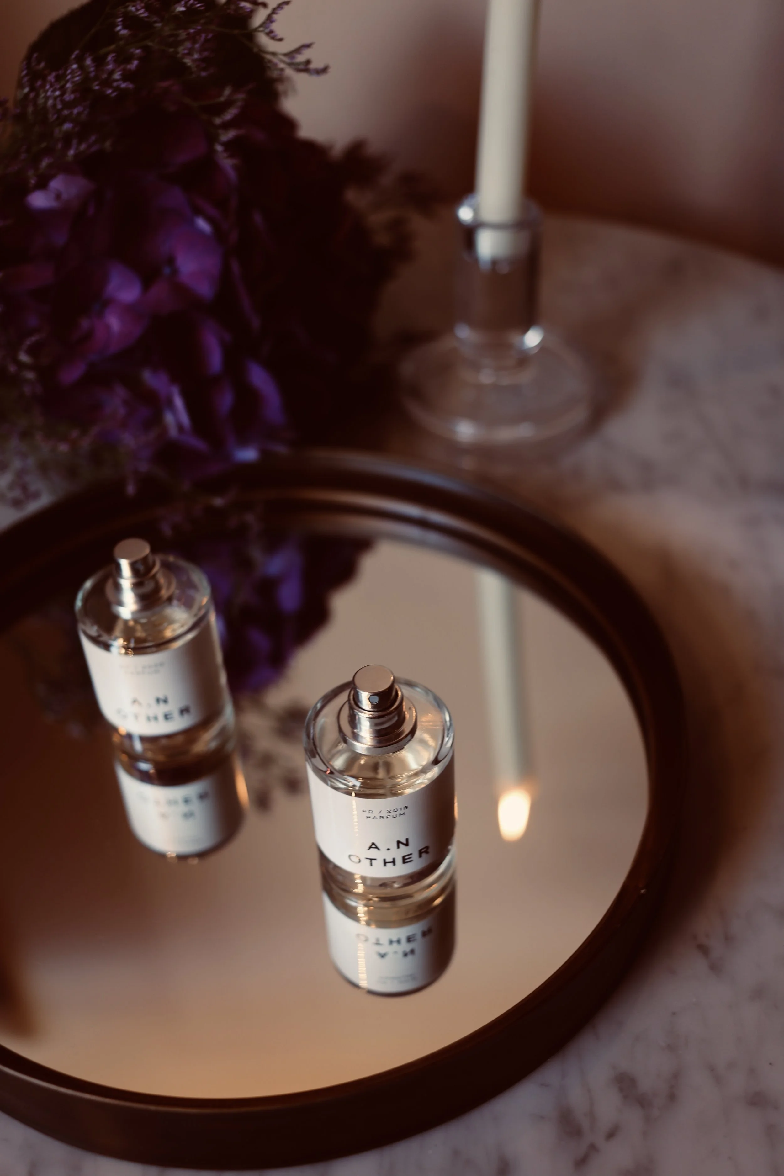 A perfume bottle labeled 'A.N Other' reflected in a round mirror on a marble surface, with purple flowers and a white candle in the background.