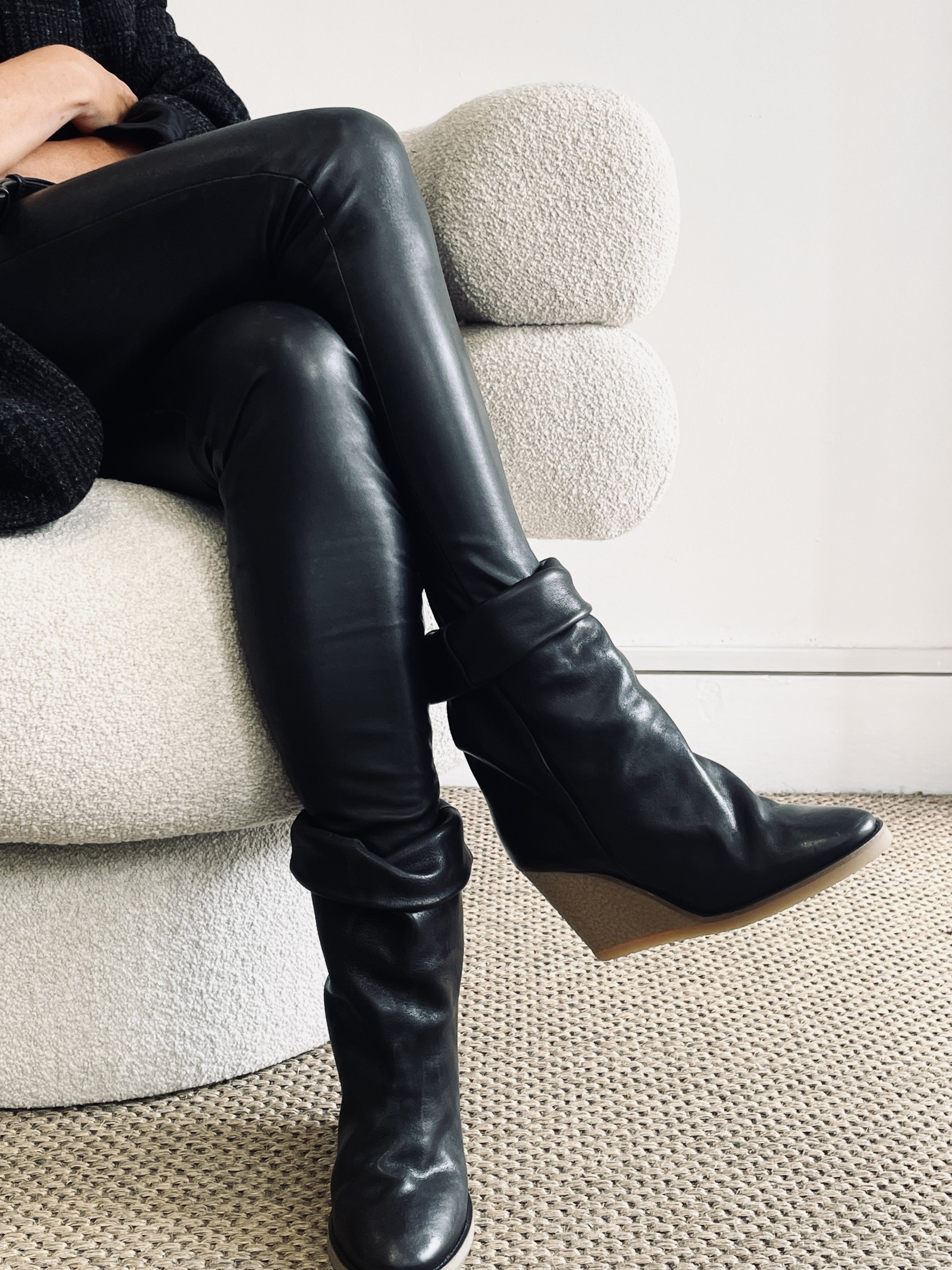 Close-up of a person sitting on a textured white sofa, wearing shiny black leather thigh-high wedge boots with a pointed toe and a curved wedge heel, with a beige carpeted floor below.
