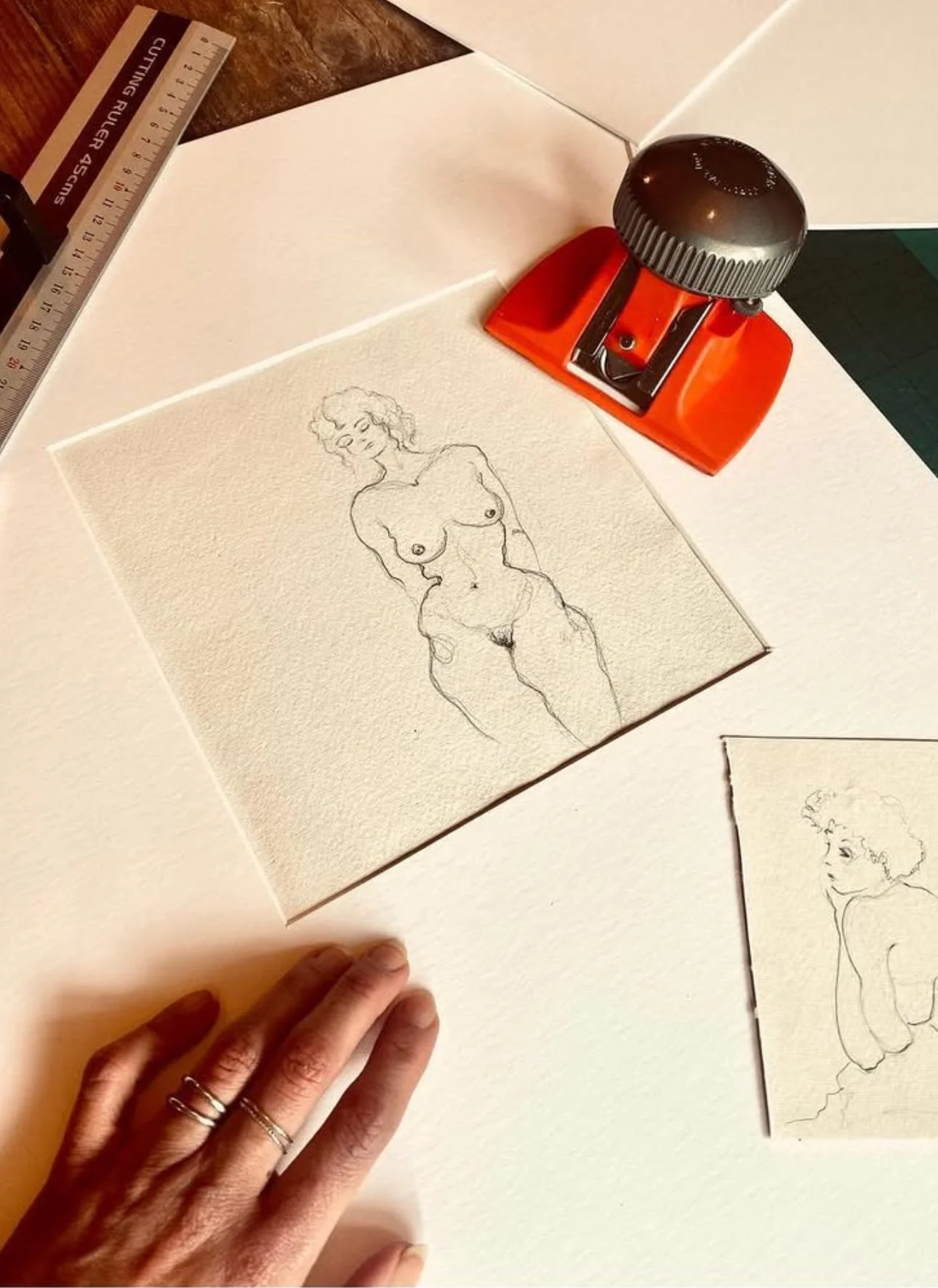 A line drawing of a nude woman with curly hair on textured paper, placed on a white surface with a hand nearby, a ruler, and a red stapler.