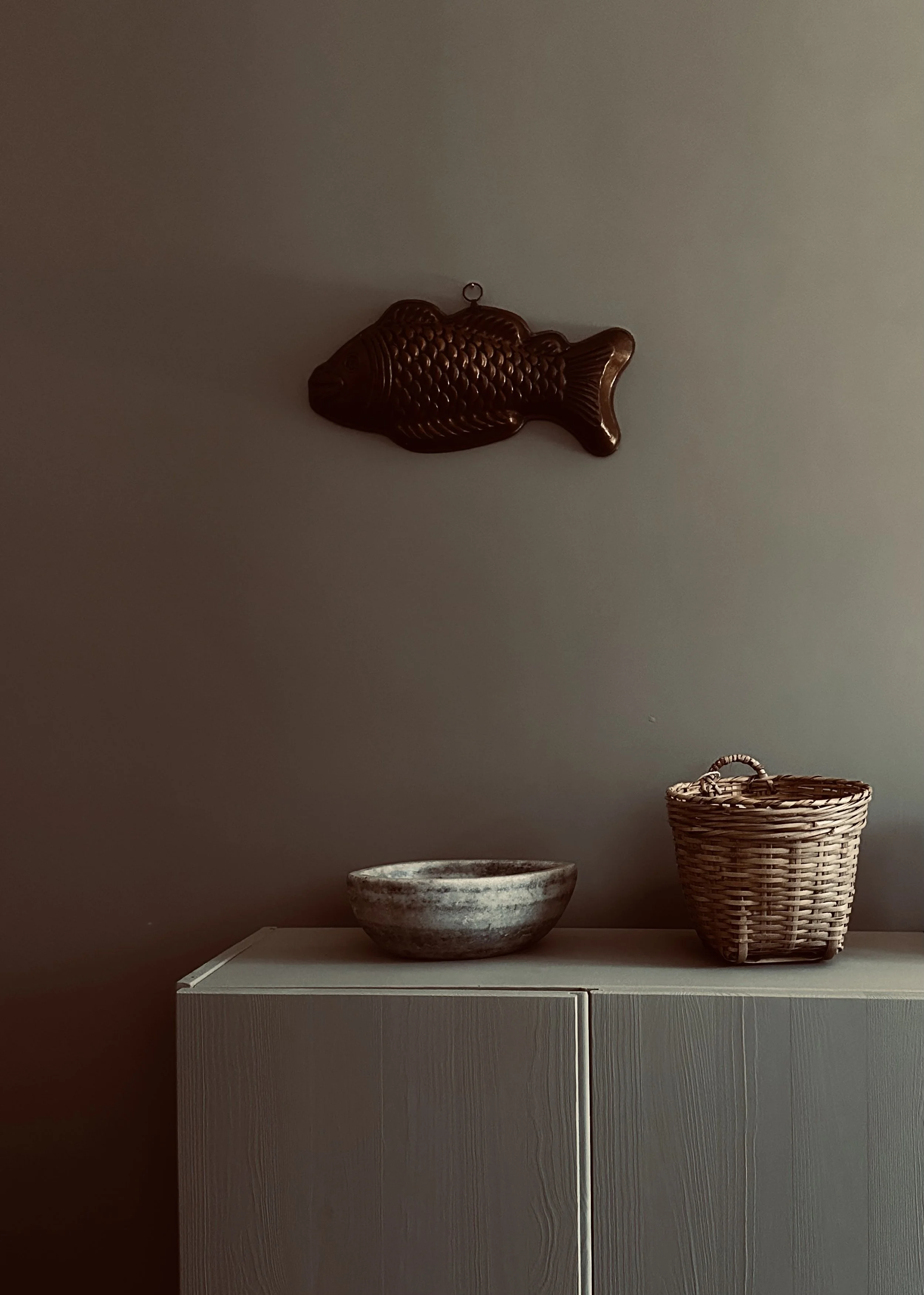 A decorative wall hanging of a fish above a cabinet, with a ceramic bowl and a wicker basket on top.