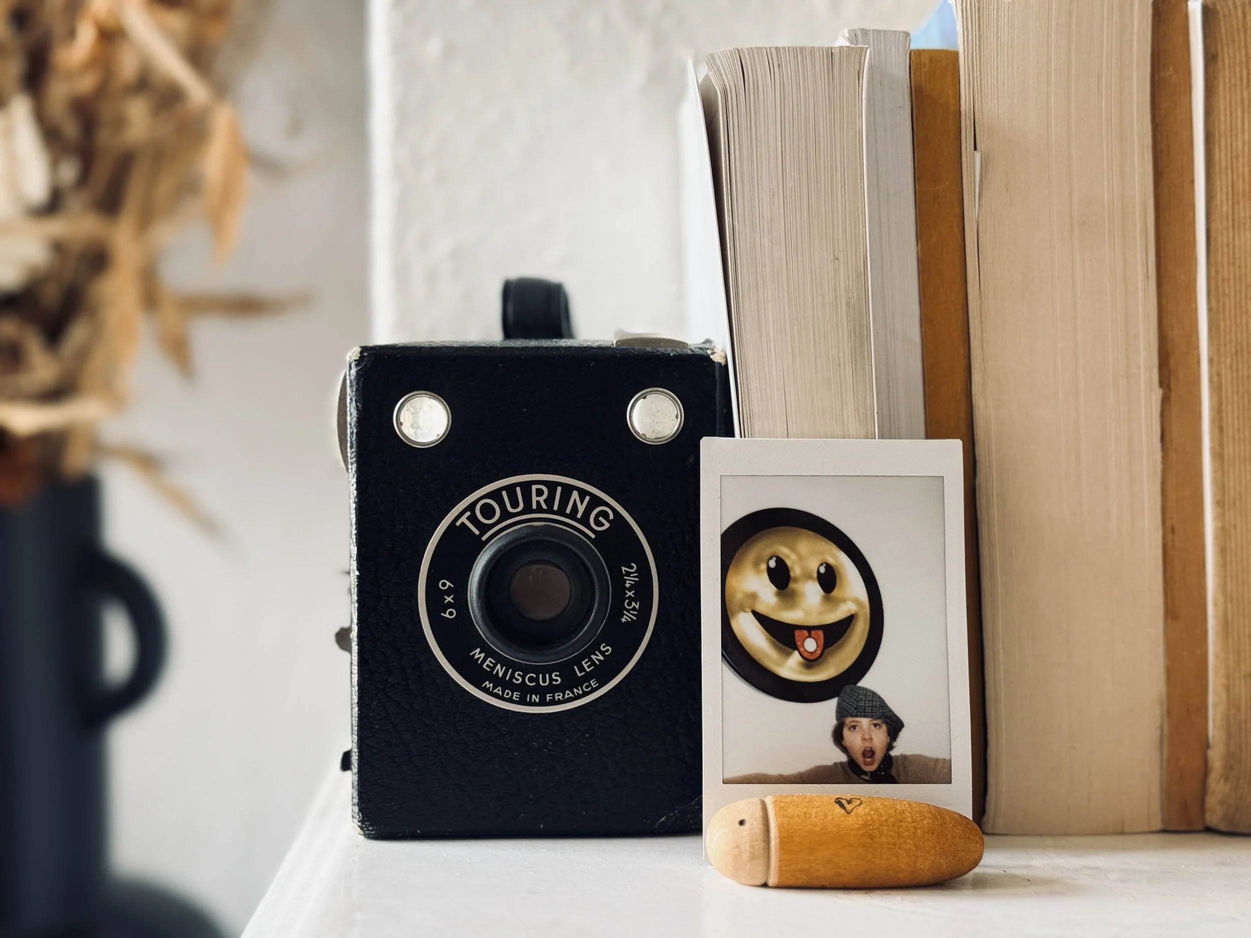 Vintage black camera, a photo with a smiley face sticker, a person with a surprised expression wearing a gray hat, and a small wooden figurine in front of books on a white surface.