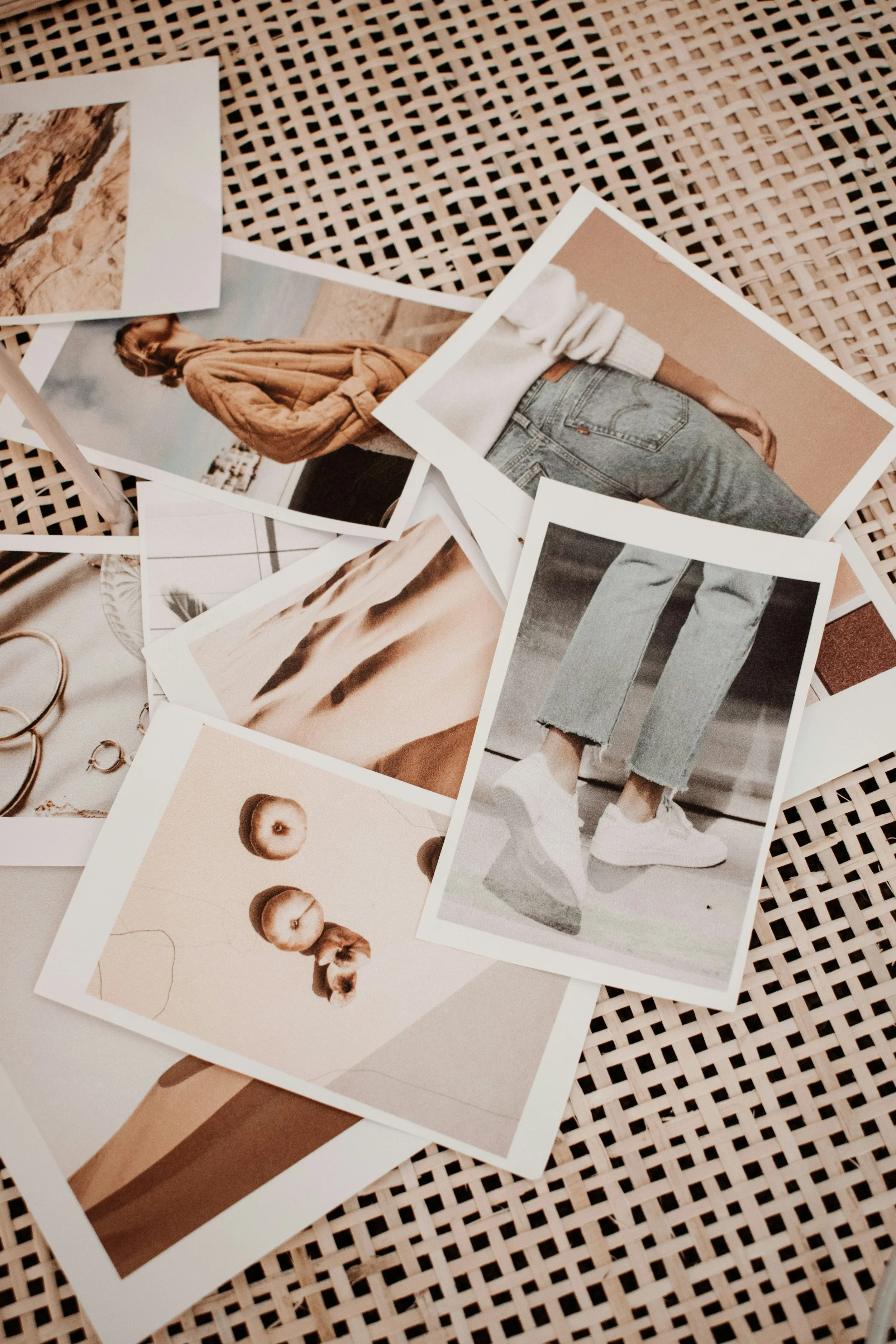 A collection of printed photographs spread out on a woven surface, including images of a woman in a tan coat, close-up of accessories, deserts, and fruits.