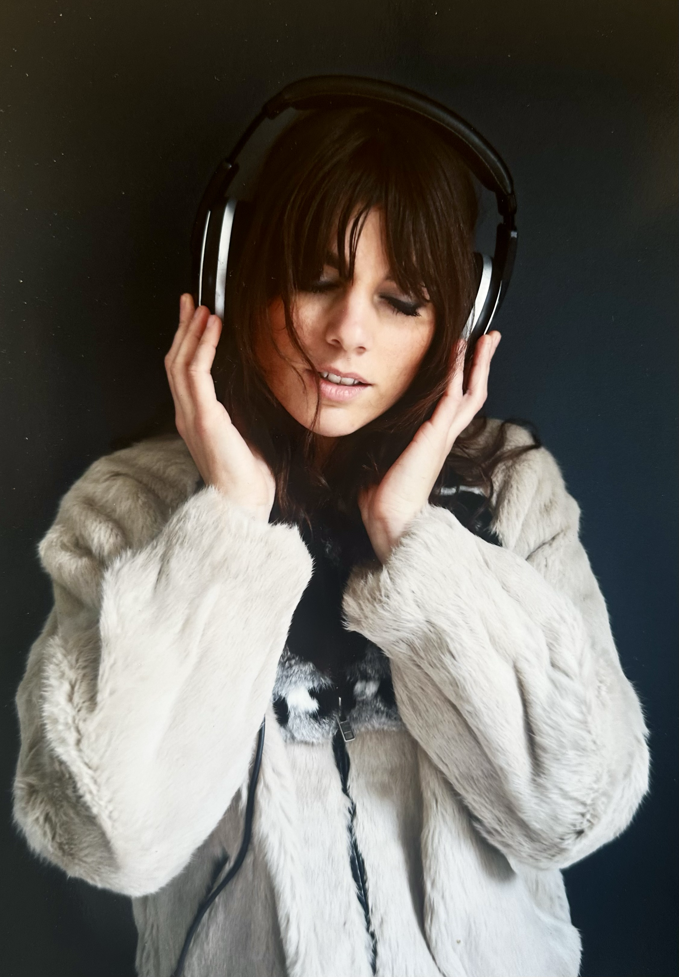 A woman with long dark hair, wearing a light fur coat, is listening to music through large black headphones with her eyes closed and hands adjusting the headphones against her ears, standing against a dark background.