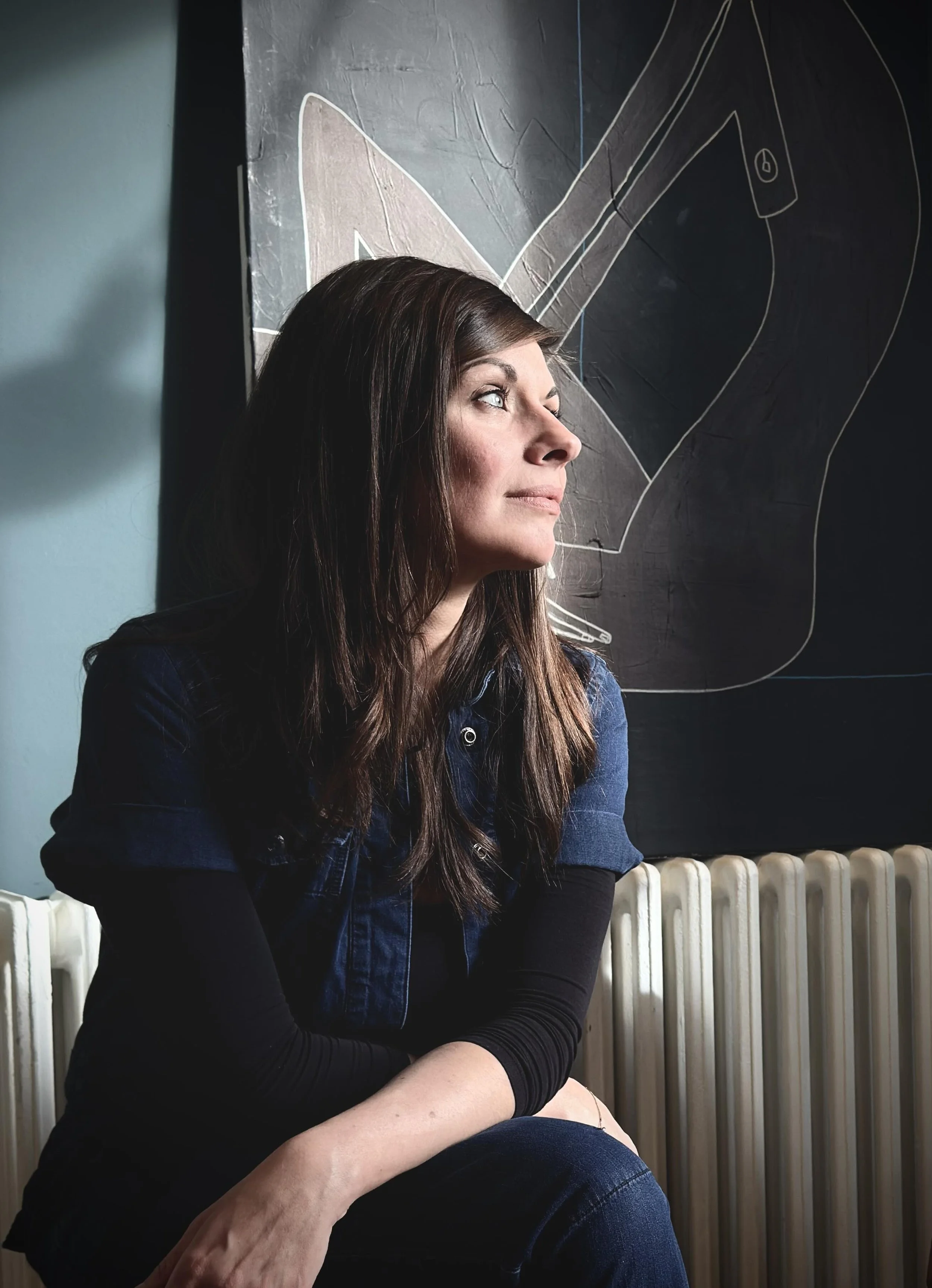 A woman sitting by a radiator, gazing thoughtfully out of the window with a chalkboard art of a face drawing visible behind her.