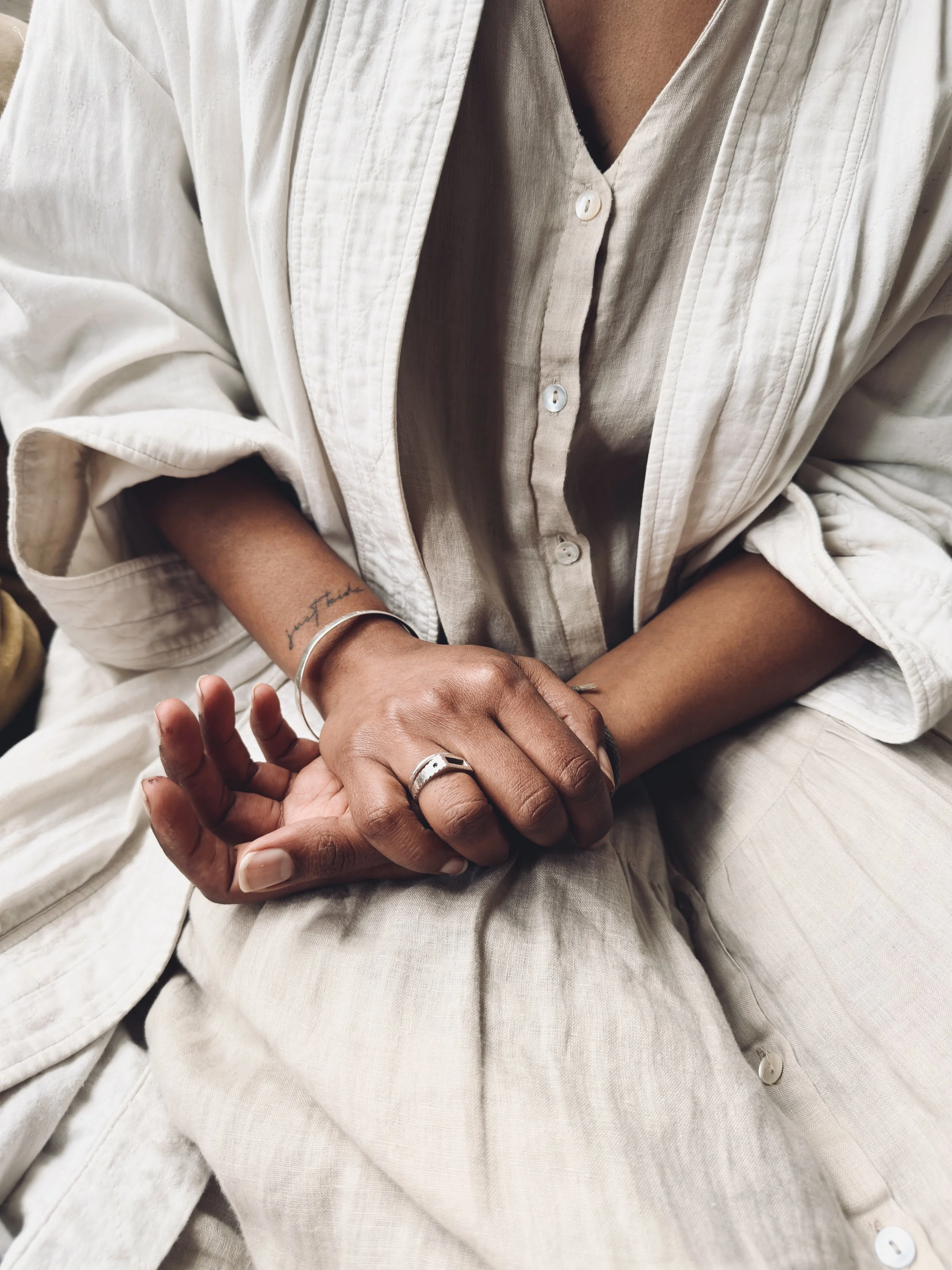 A person wearing a beige button-up shirt sitting with their hands clasped, showing rings and a bracelet.