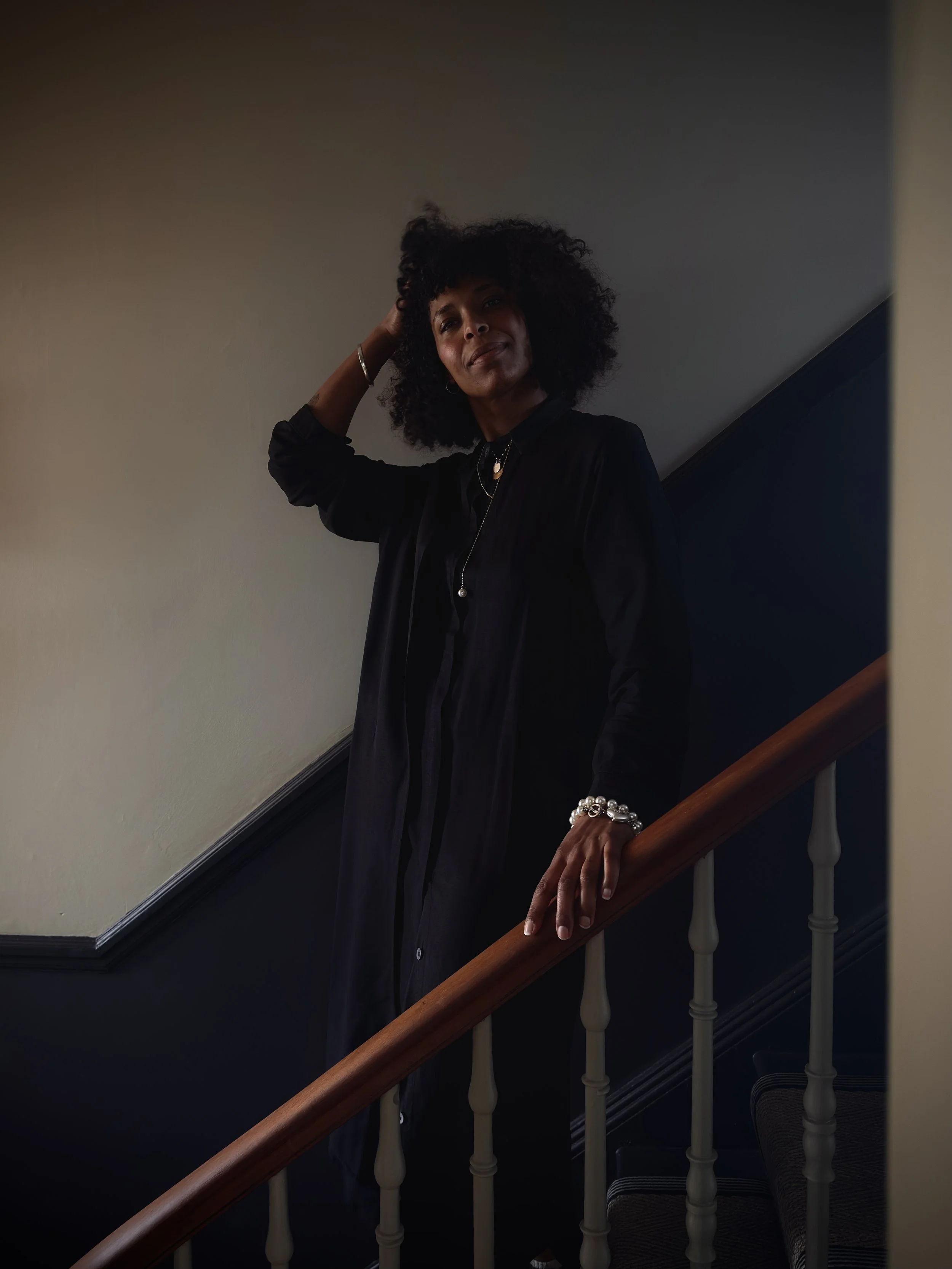A woman with curly hair standing on a staircase, touching her head with one hand and resting her other hand on the railing, wearing dark clothing and jewelry.