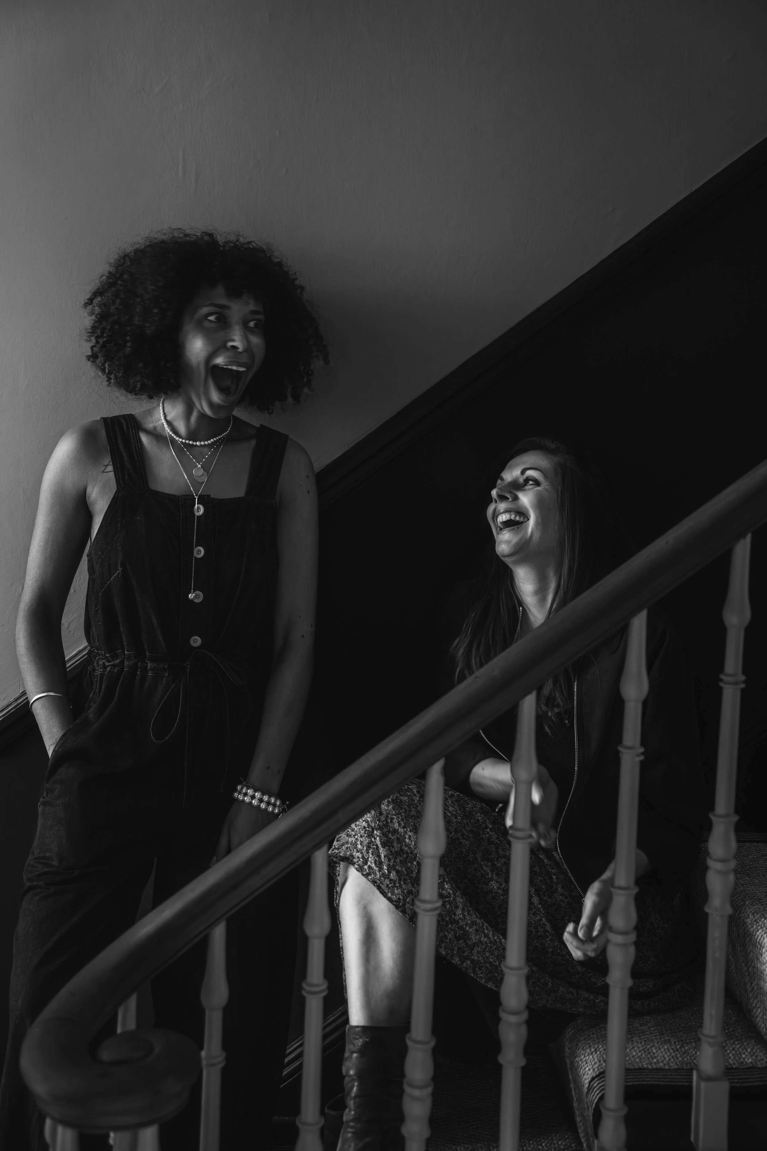 Two women laughing and talking on a staircase indoors.