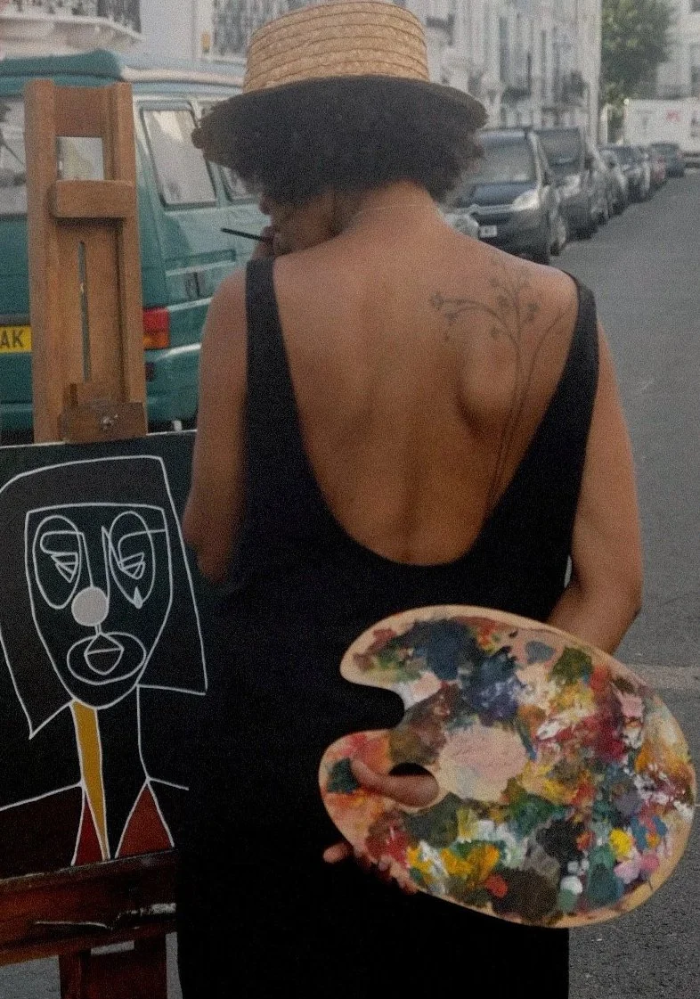A woman wearing a straw hat and black dress with open back, holding a painter's palette, standing on a street with parked cars and a chalk art of a face on an easel.