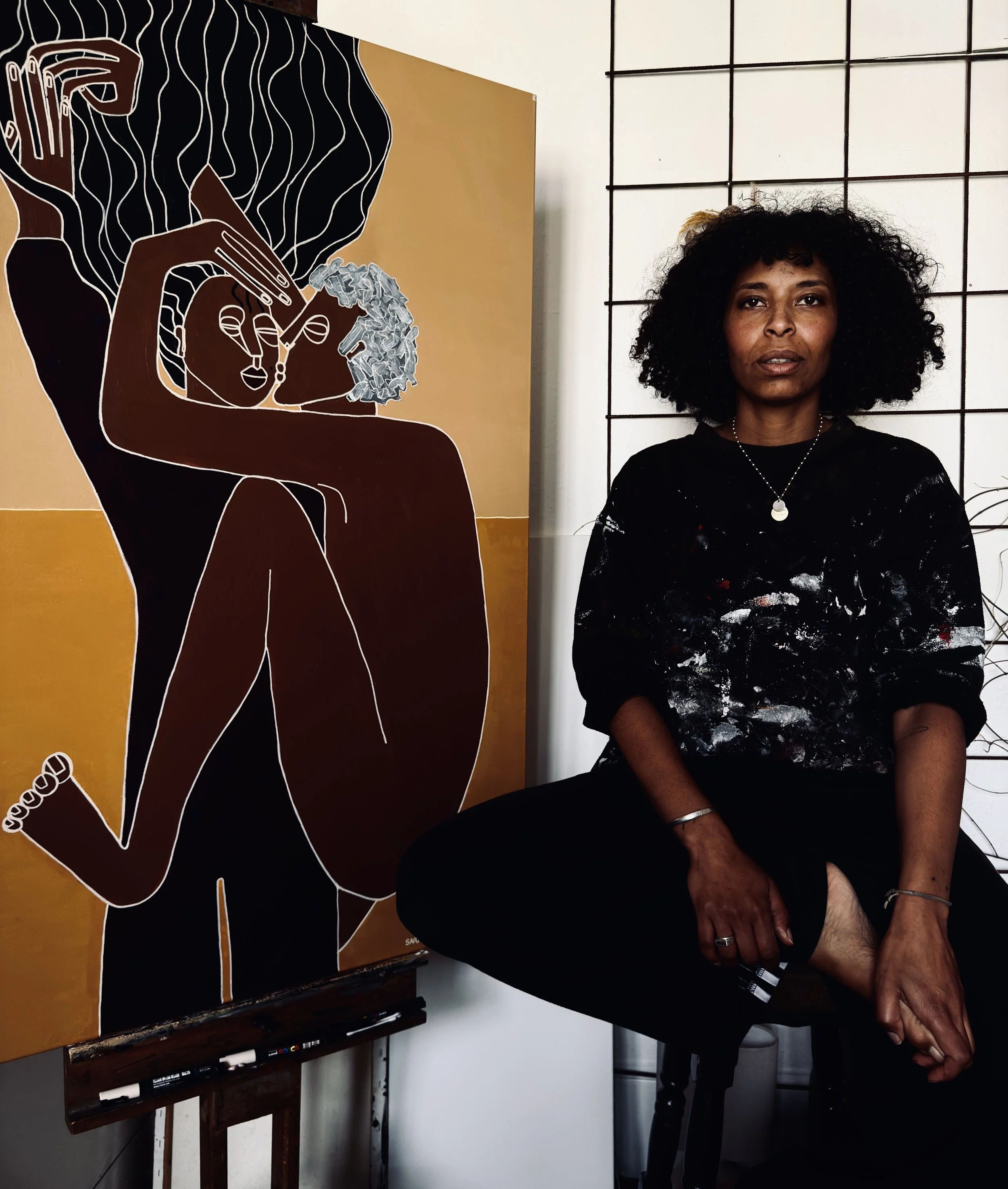 An artist with curly hair sitting next to an abstract painting of a woman and a child. The paintings and artist are in a studio setting.