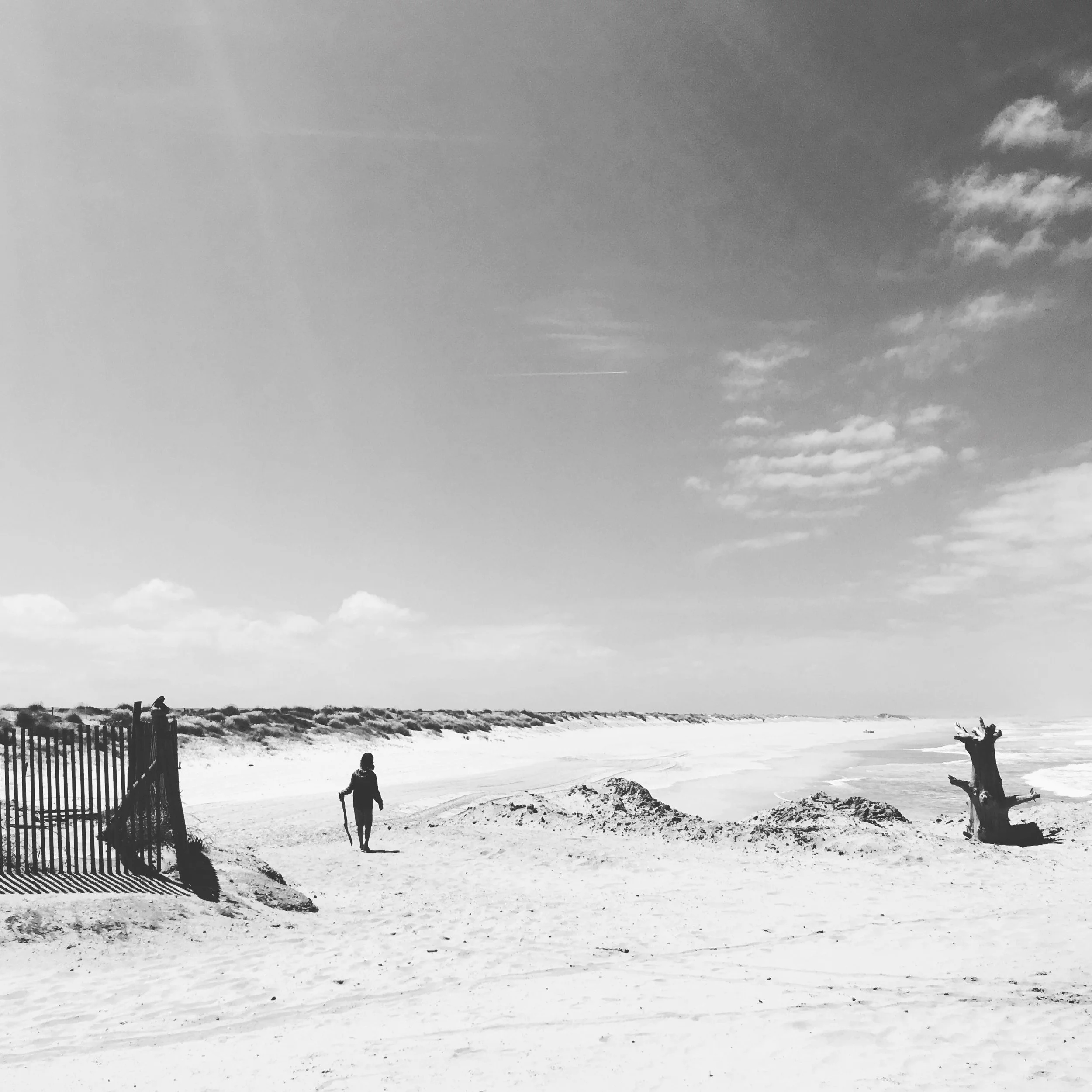 A black and white photo of a person walking along a sandy beach with a walking stick, near a fence and a piece of driftwood, with a vast sky and distant shoreline.