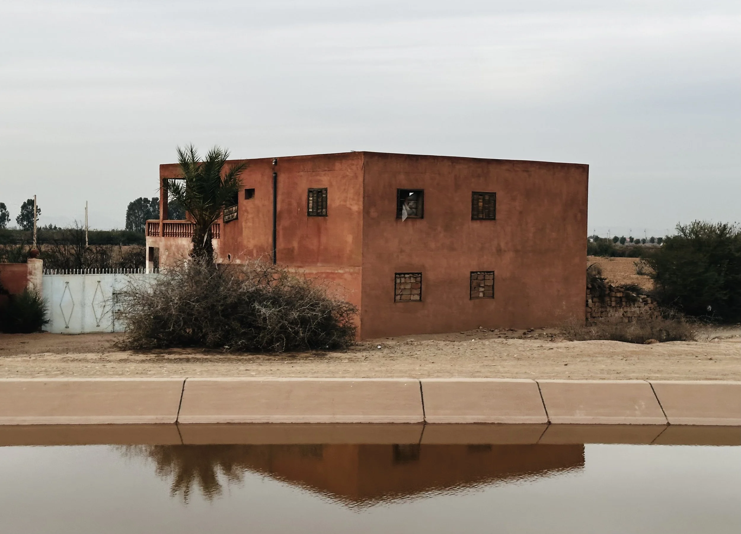 A two-story, earth-toned house with small, barred windows, a palm tree nearby, and dry desert surroundings reflected in a water feature in the foreground.