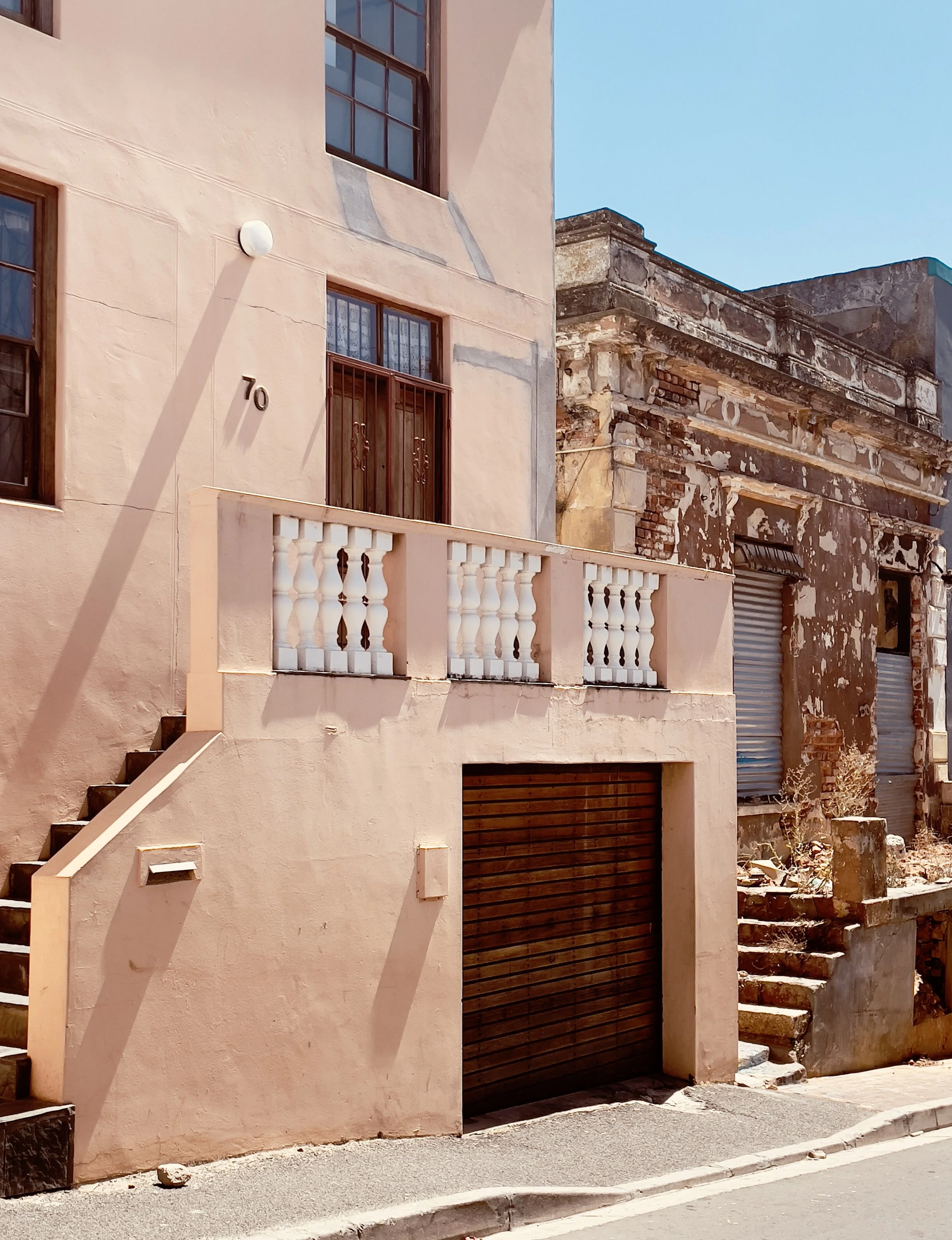 A pink building with a balcony, staircase, and garage door, adjacent to a dilapidated structure with peeling paint and closed shutters, sunny street scene.