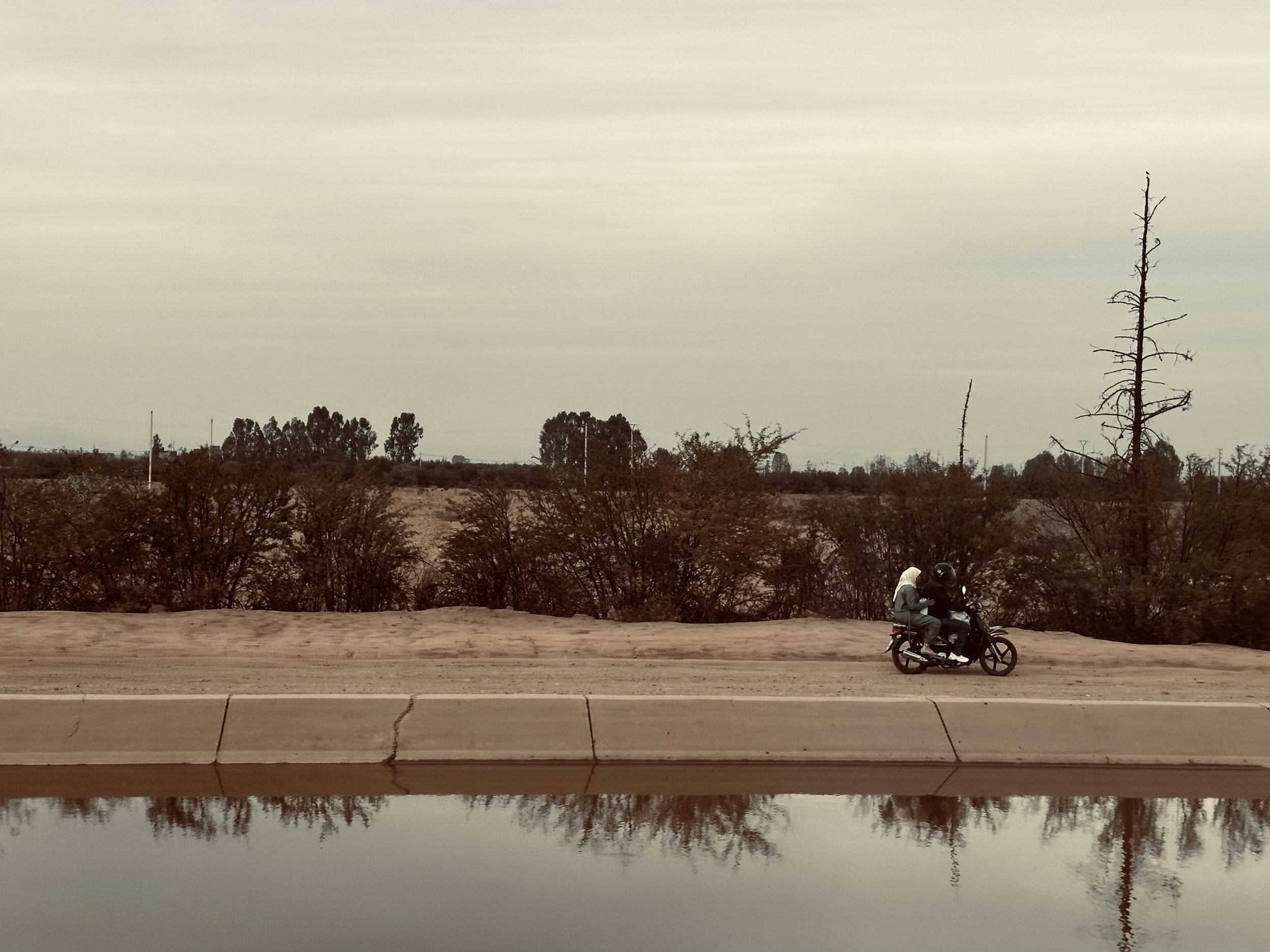 Two people riding a motorcycle along a roadside with dry trees and bushes, and a reflective water surface in the foreground under an overcast sky.