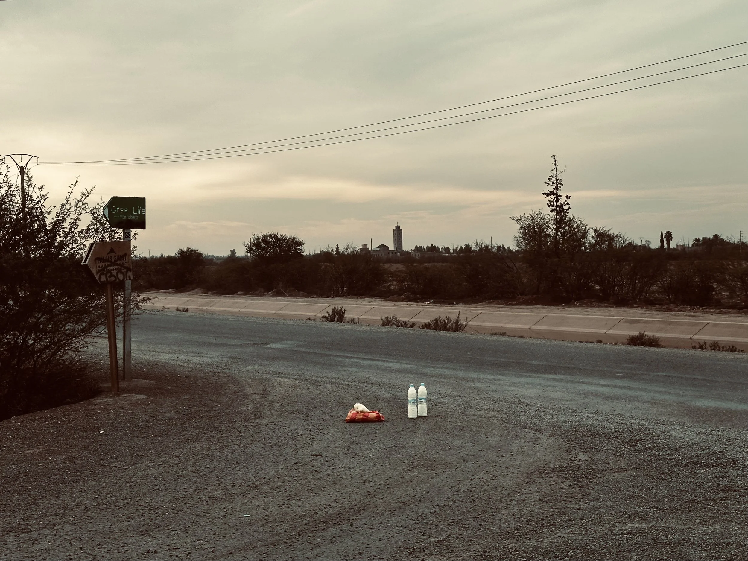 Two bottles of water and a red plastic bag lying on the ground beside a deserted road in a desert landscape with shrubs and sparse trees, under a cloudy sky.