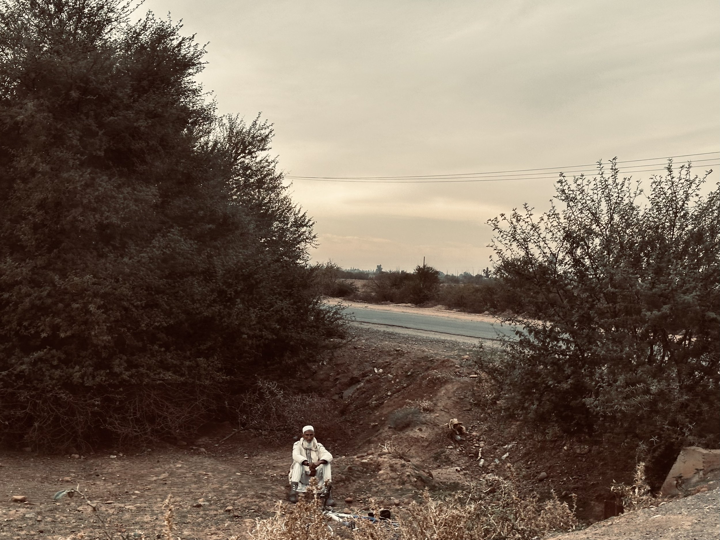 A man sitting on the ground near a dirt road with bushes and trees on either side and power lines in the background under a cloudy sky.
