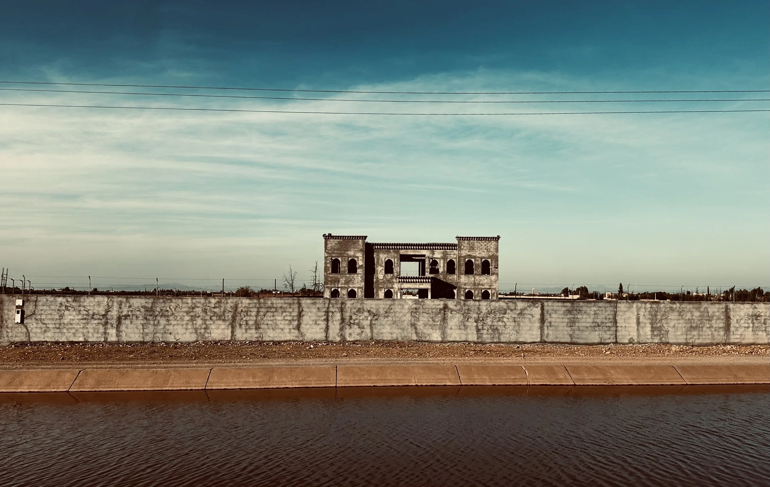 An unfinished or abandoned two-story building with arched windows, surrounded by a concrete wall and a body of water in the foreground, under a blue sky with light clouds.