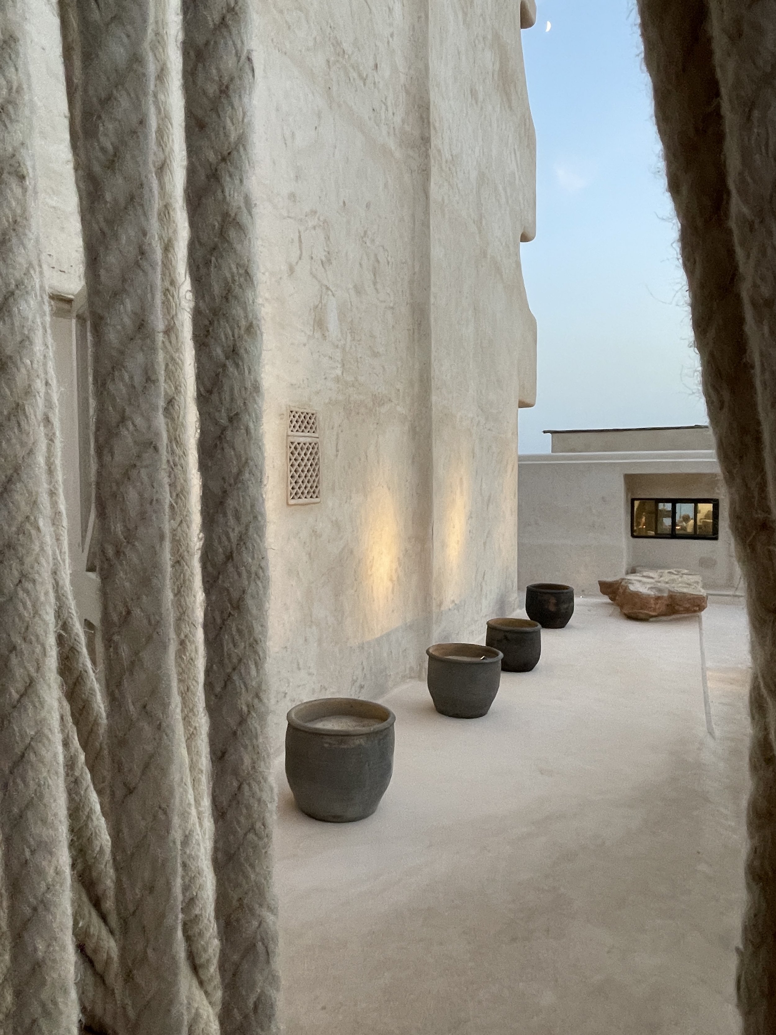 A view through a window of a minimalist courtyard with a textured white wall, five decorative pots, and a large rock, with a small window and the sky in the background.