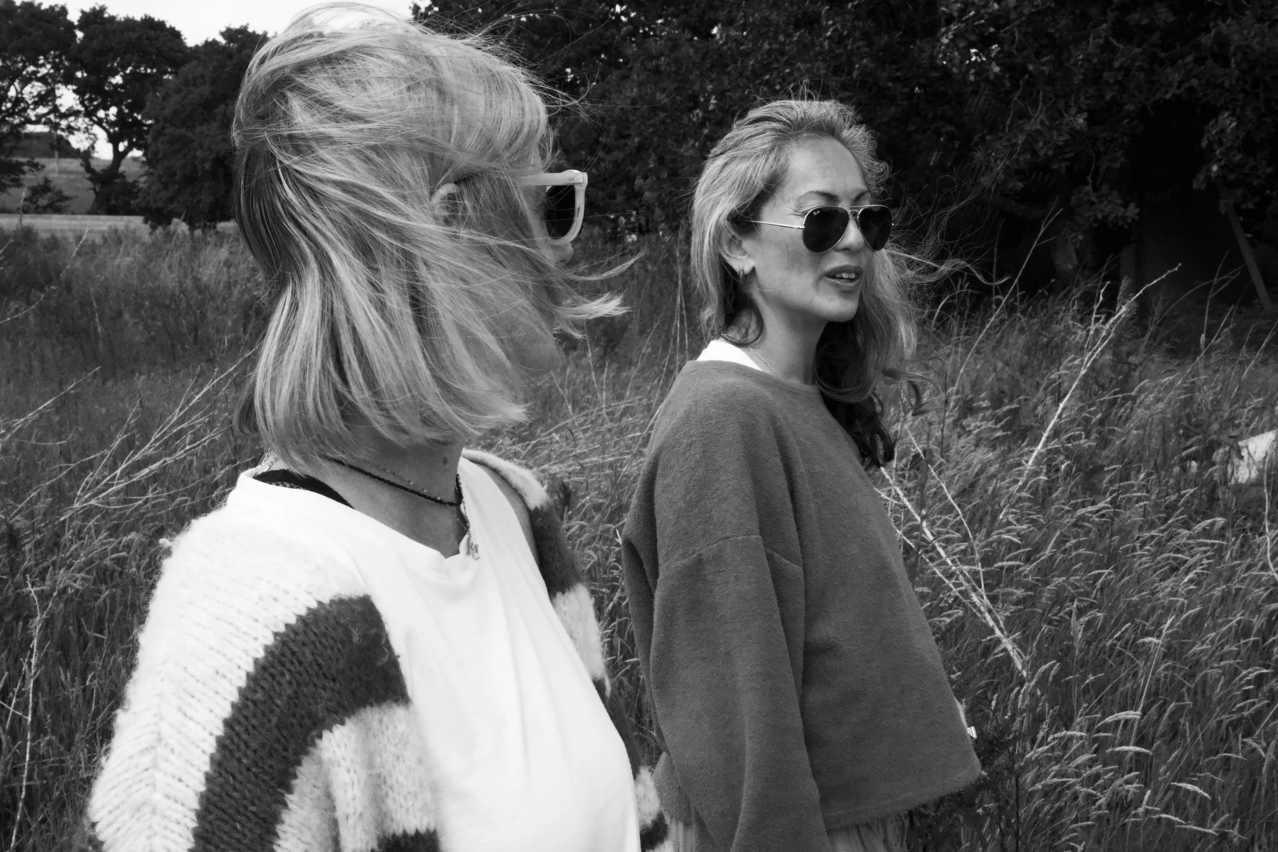 Two women wearing sunglasses standing outdoors in a grassy field with trees in the background.