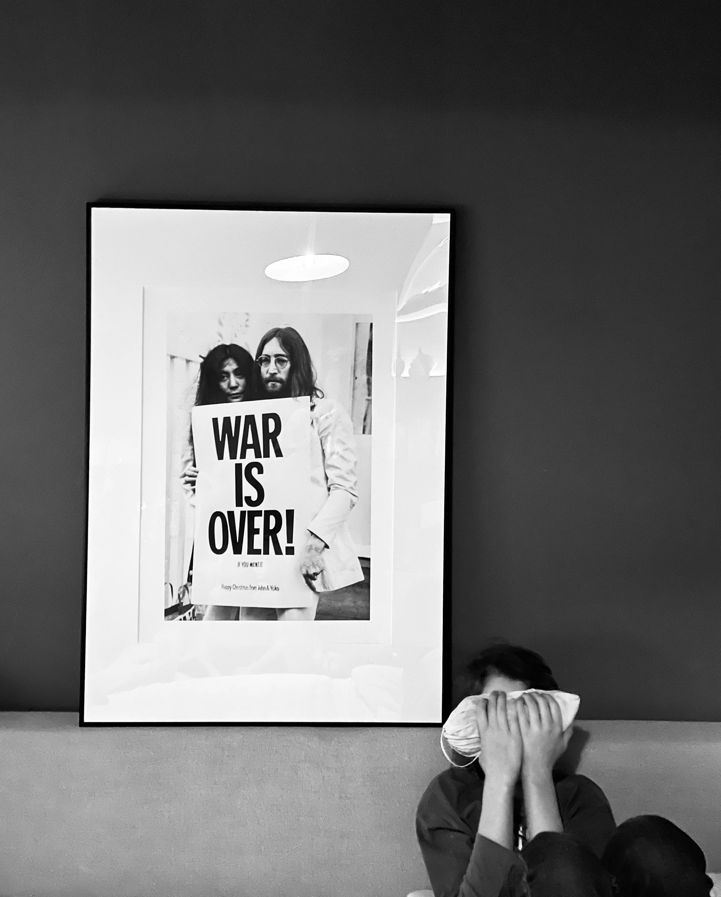A person covering their face with a cloth sitting in front of a framed black-and-white photo of two people holding a sign that reads 'WAR IS OVER!'
