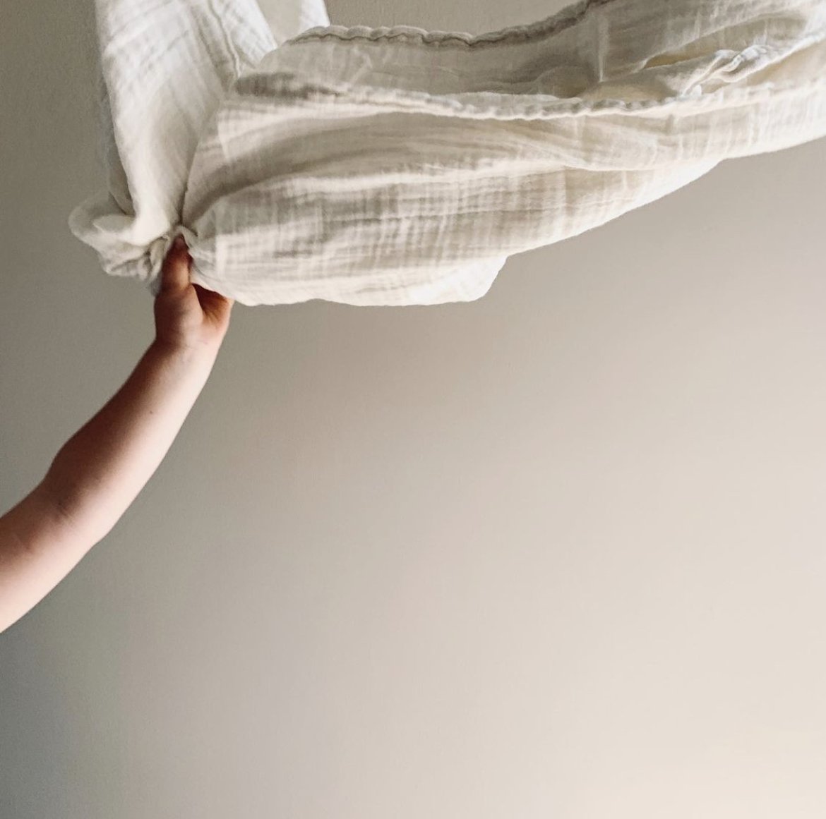 A person's hand gently holding the corner of a cream-colored, striped bedsheet or blanket against a plain, off-white wall.