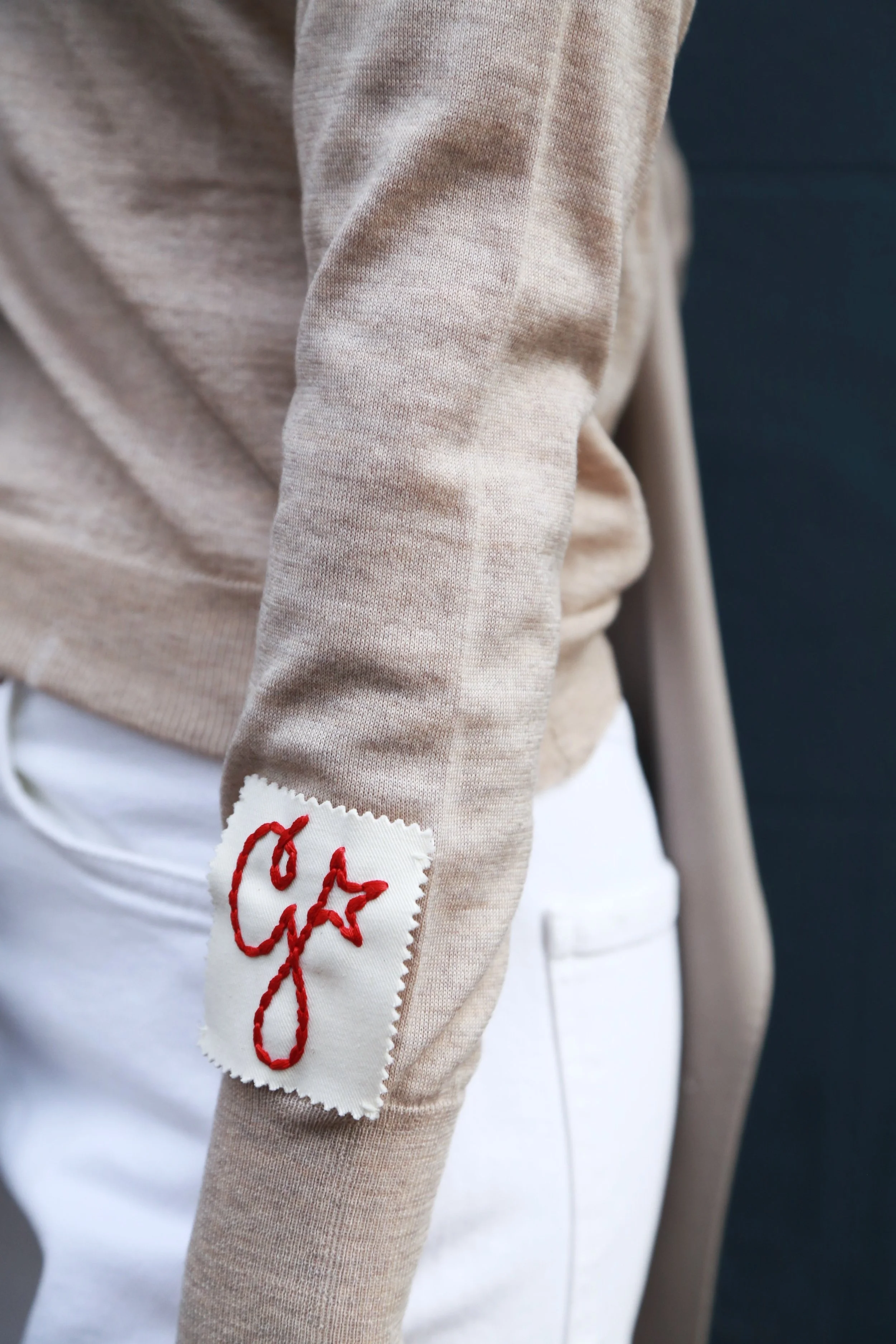 Close-up of a beige sweatshirt with a white patch on the sleeve that has red embroidery of a signature or writing.