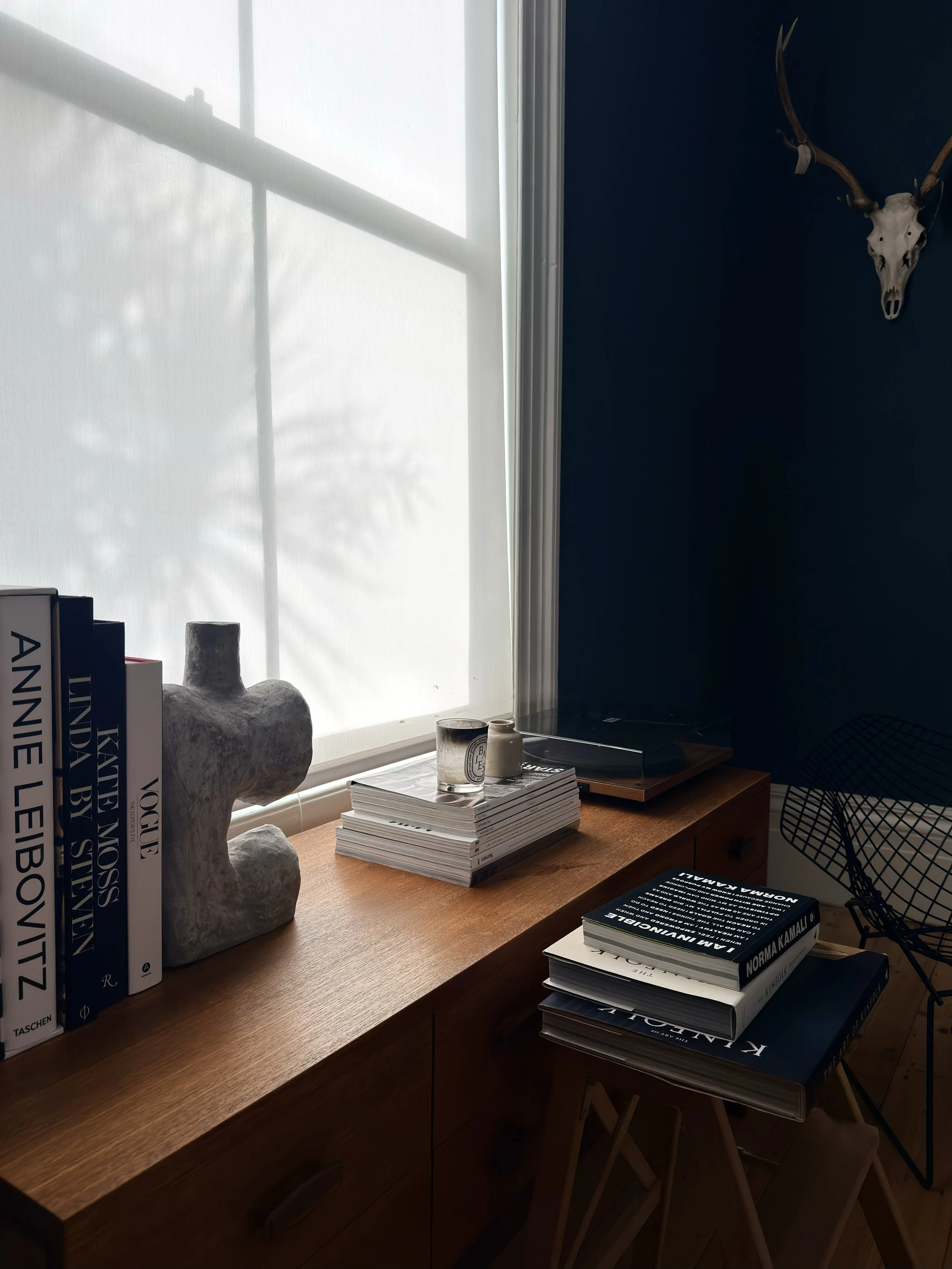 Sunlight streaming through a window onto a wooden desk with books, a stone sculpture, and small candles, with a deer skull mounted on a dark blue wall.