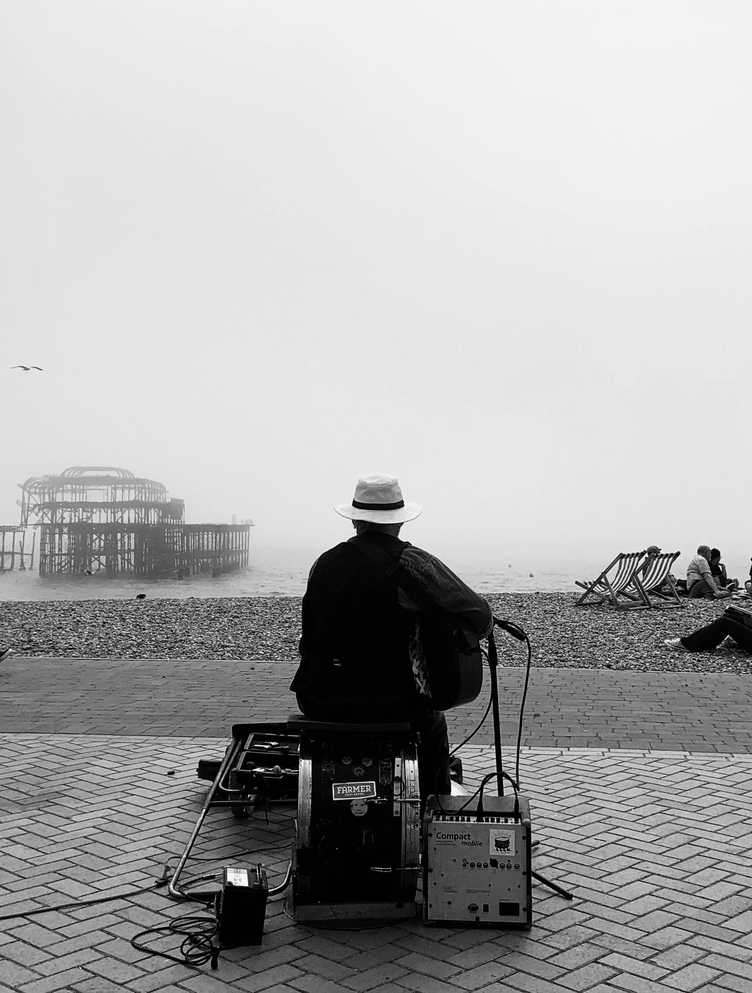 A man with a hat playing guitar on a beachside promenade with the pier visible in the foggy background, some people sitting on the beach and lounge chairs nearby.